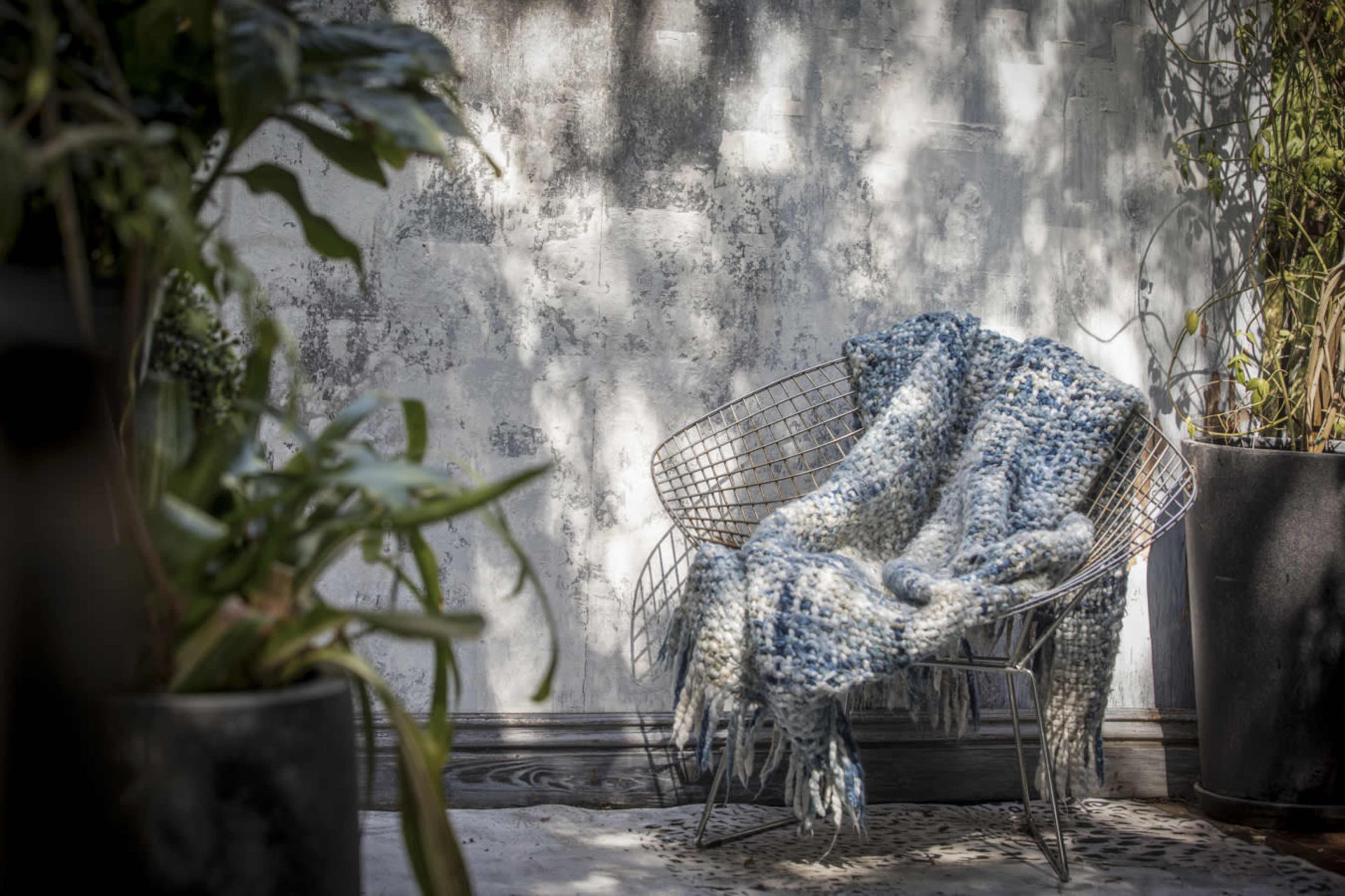 A knitted blue blanket is draped over a wire chair in a serene outdoor setting with potted plants nearby.