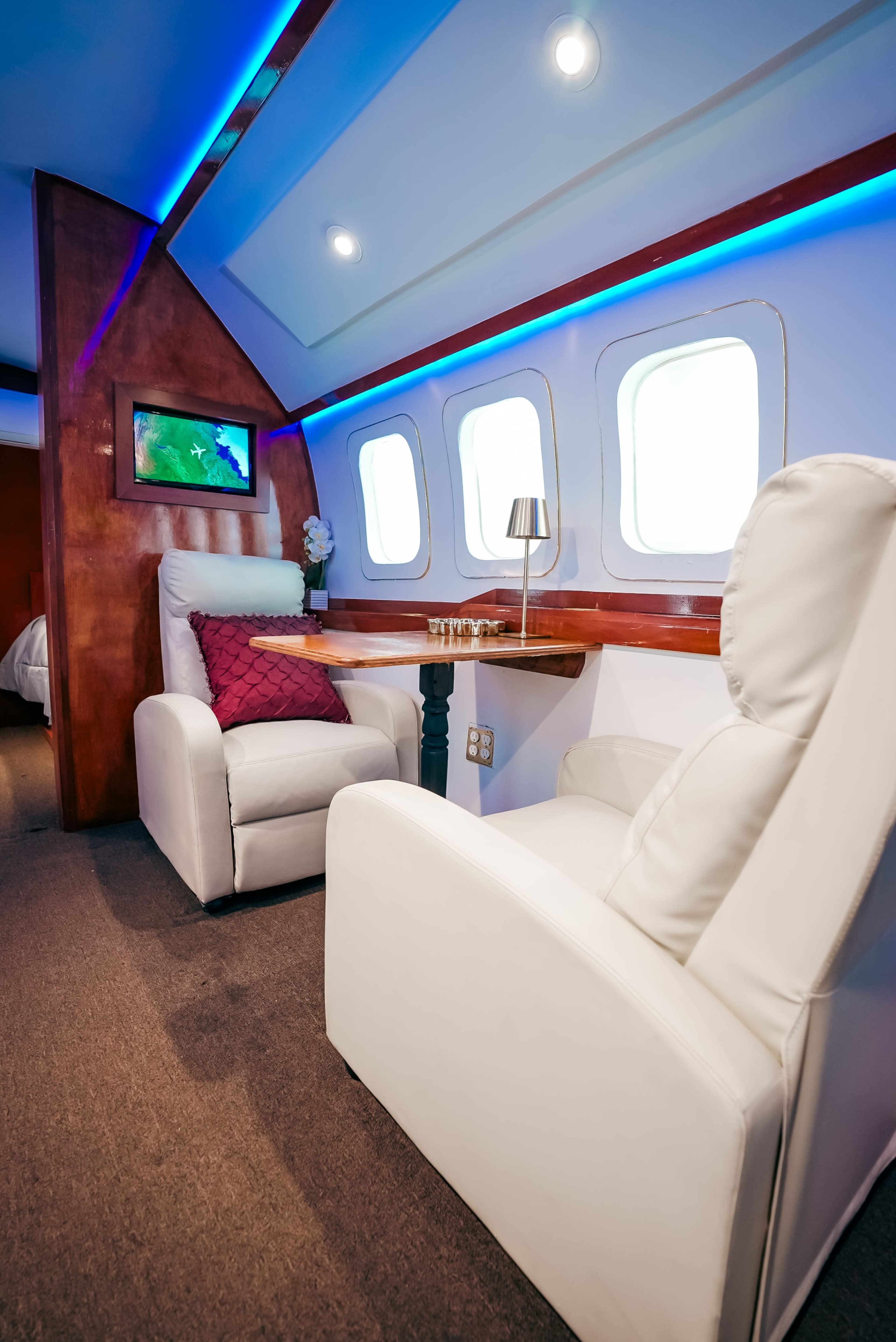 The image depicts a modern interior of a room designed to resemble an airplane cabin, featuring two white chairs, large windows, and ambient lighting.