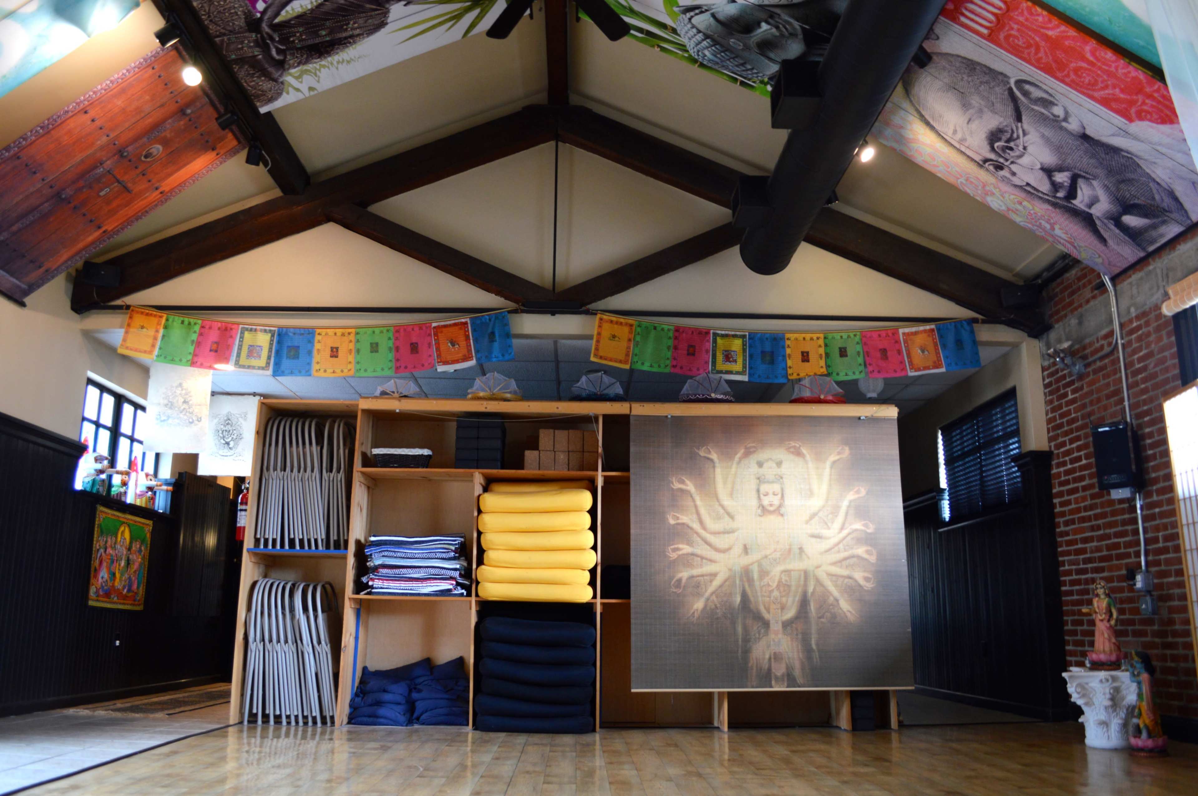 An interior space with wooden beams, colorful prayer flags hanging from the ceiling, and a large mural of a figure with multiple arms, alongside neatly stacked yoga mats and blankets on a wooden shelf.