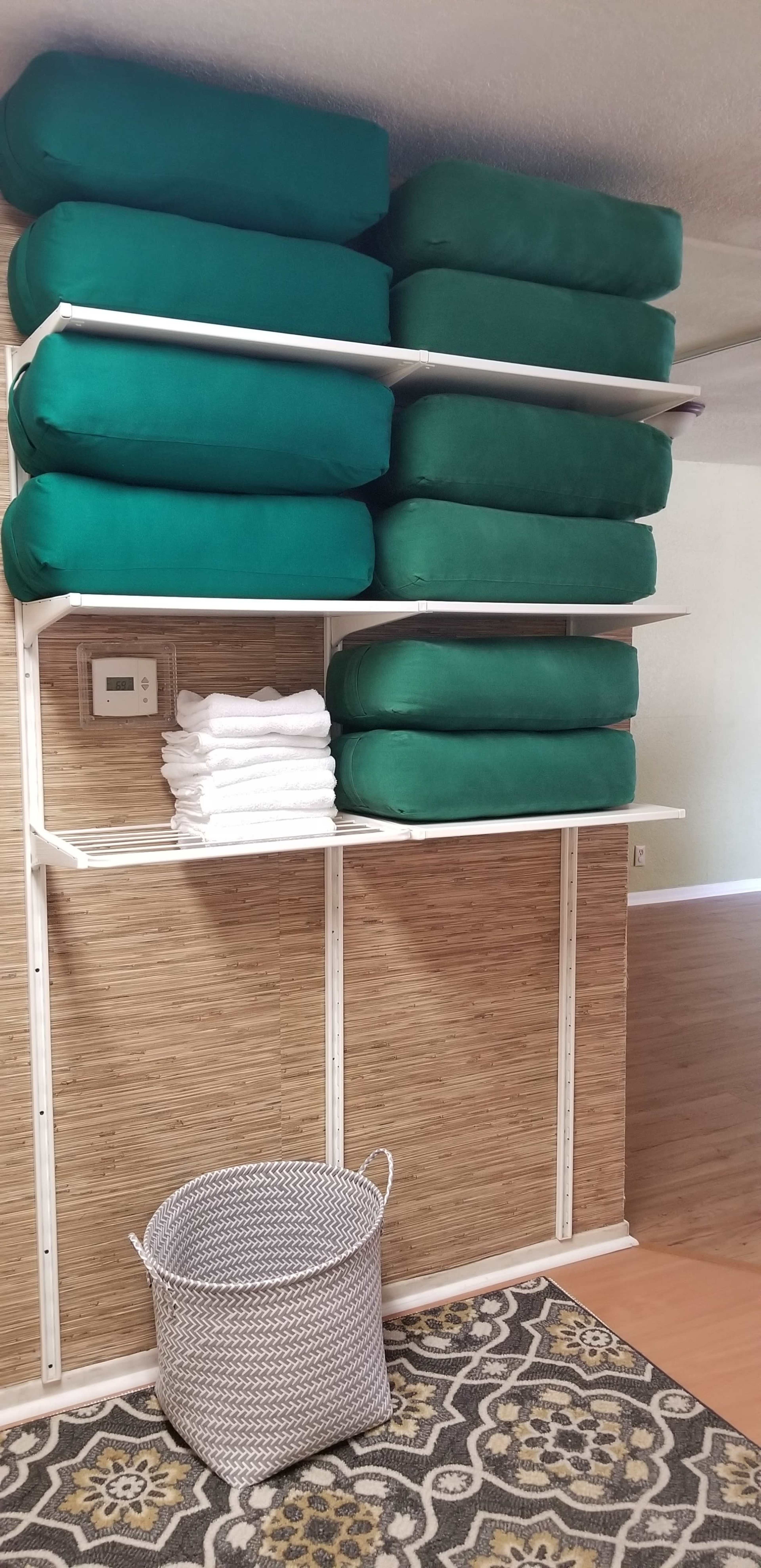 A neatly organized storage area with stacked green cushions on shelves, folded white towels, and a wire basket on the floor.