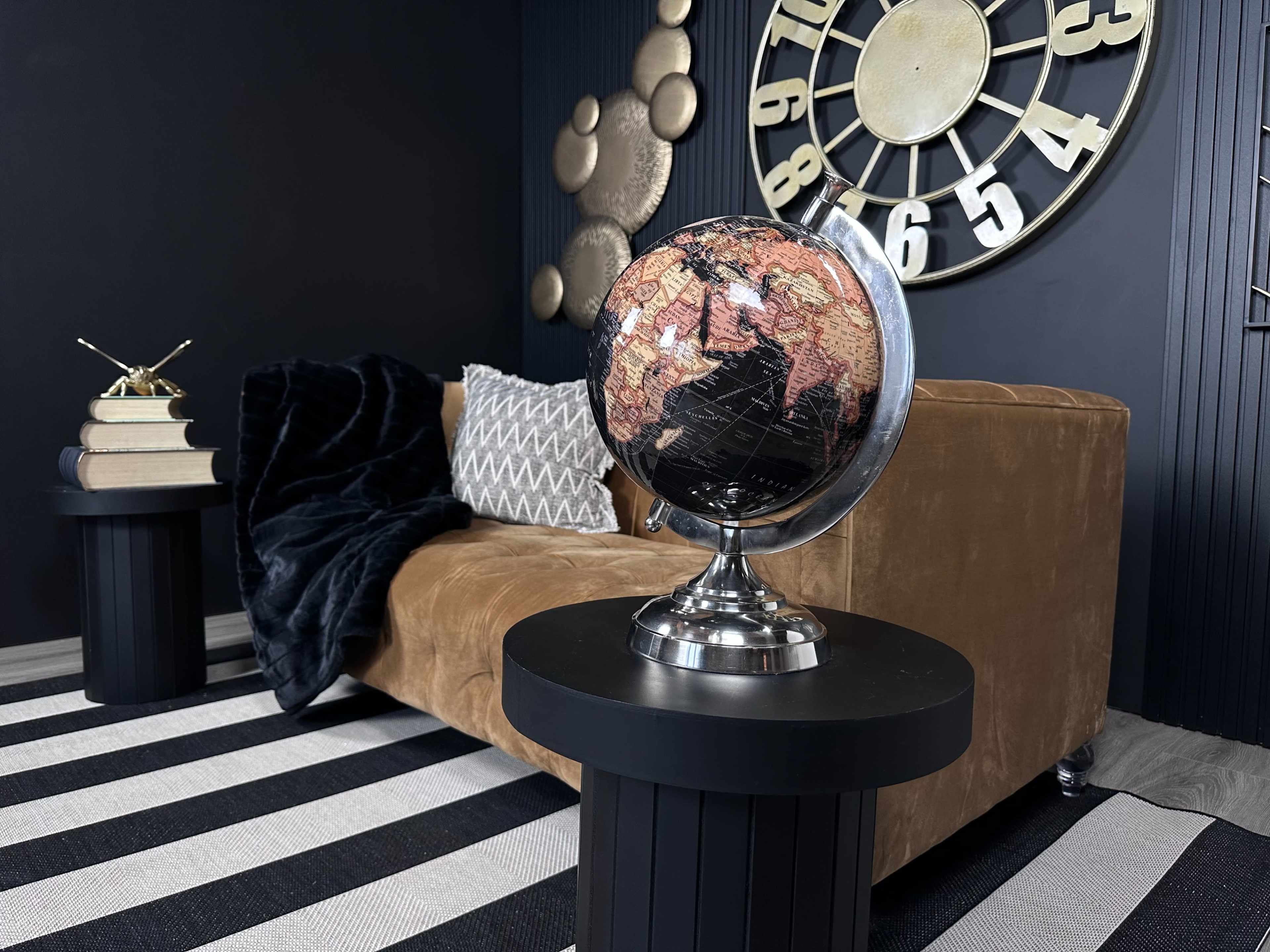 A globe on a black pedestal in front of a brown couch, complemented by decorative pillows and a large wall clock in a modern interior setting.