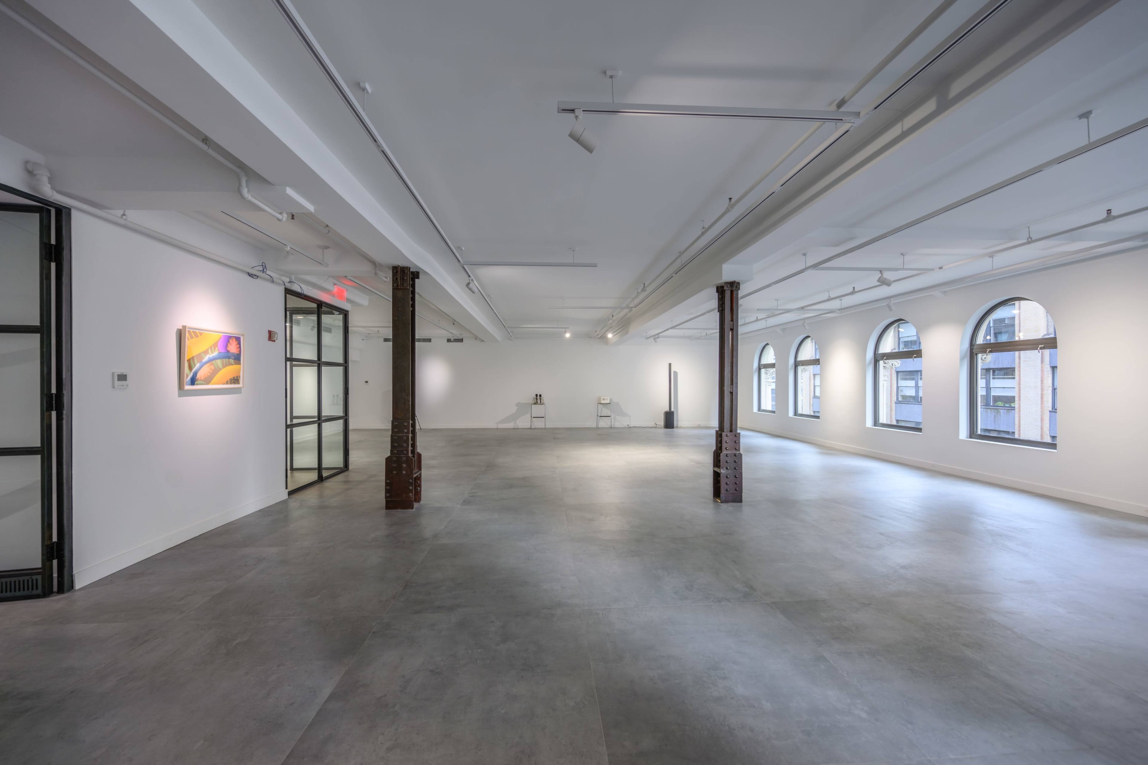 The image shows a spacious, empty room with large windows, polished gray flooring, and minimalist decor, including a painting on the wall and two chairs.