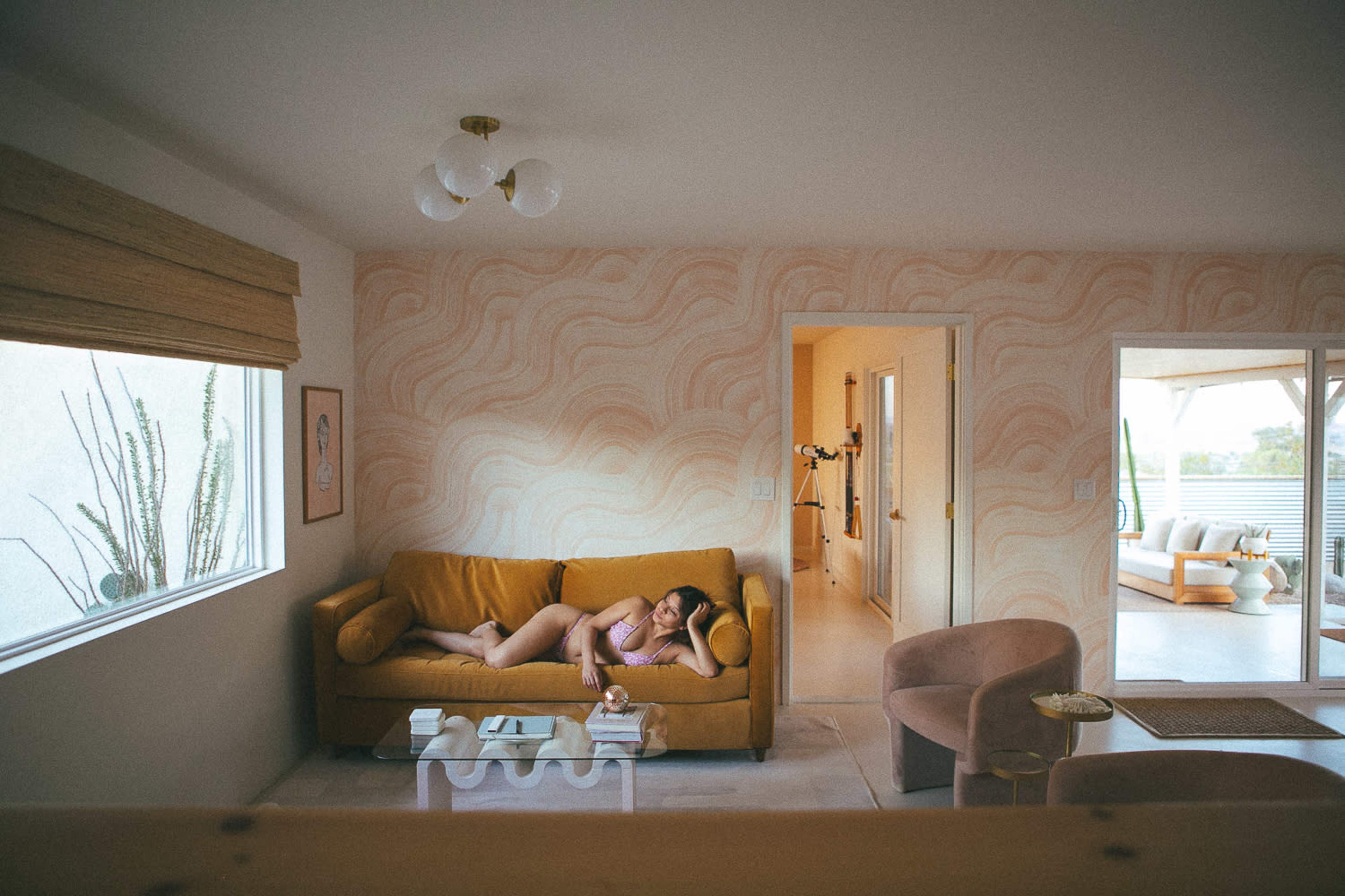 A person lies on a mustard-colored couch in a softly lit room with light-colored walls and large windows.