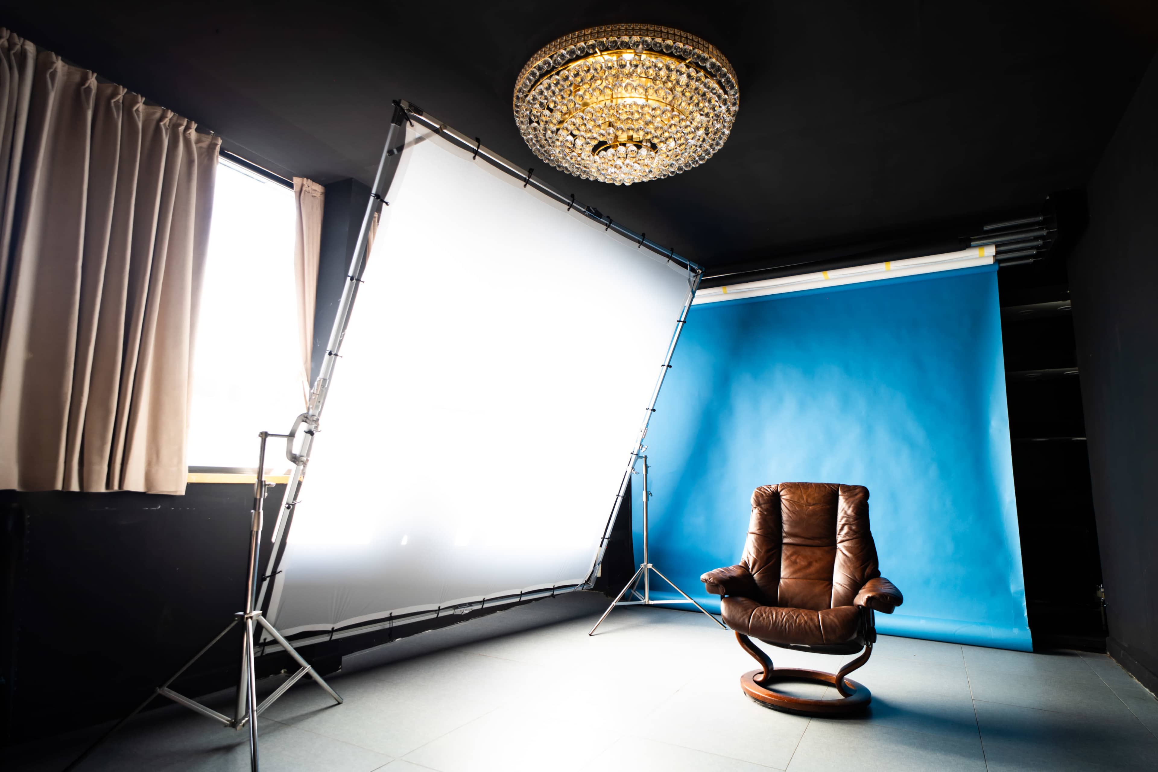 A brown leather chair is positioned in a brightly lit photography studio featuring a blue backdrop and a large chandelier.