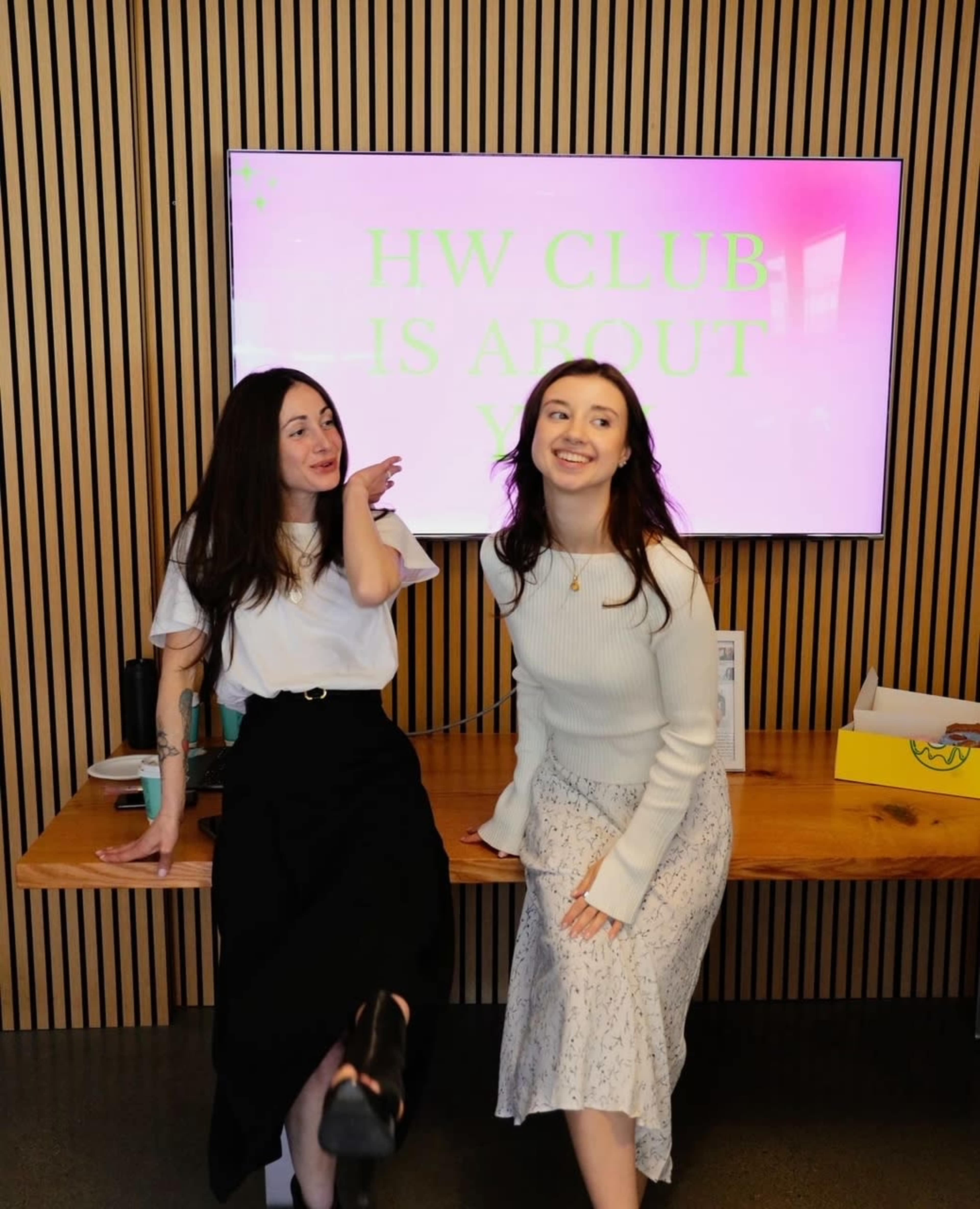 Two women pose in front of a monitor displaying the text "HW CLUB IS ABOUT YOU" in a modern indoor setting.