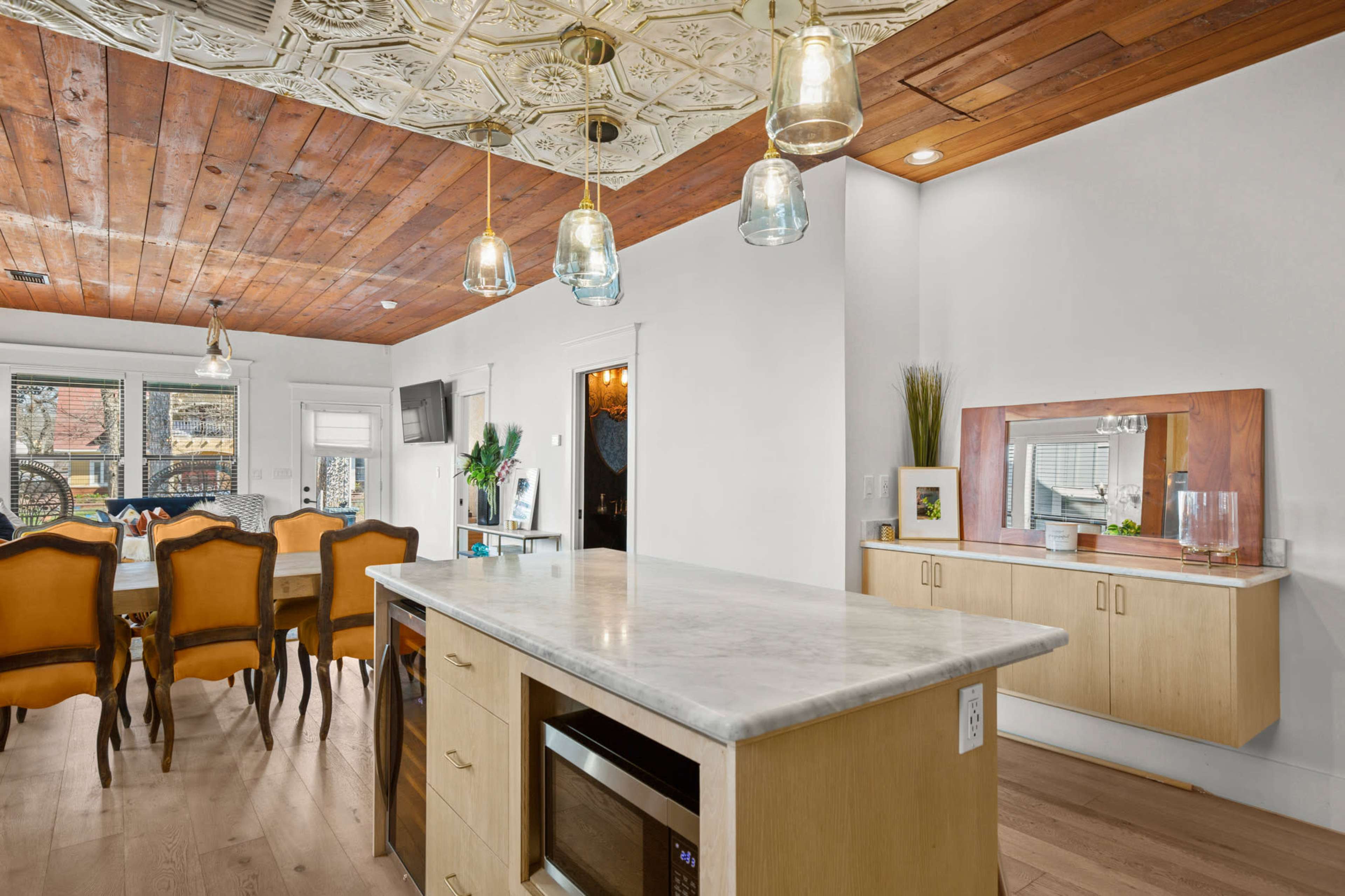 The image shows a modern kitchen with a large island featuring a marble countertop, pendant lights overhead, and a dining area in the background with a wooden ceiling and a table set for meals.