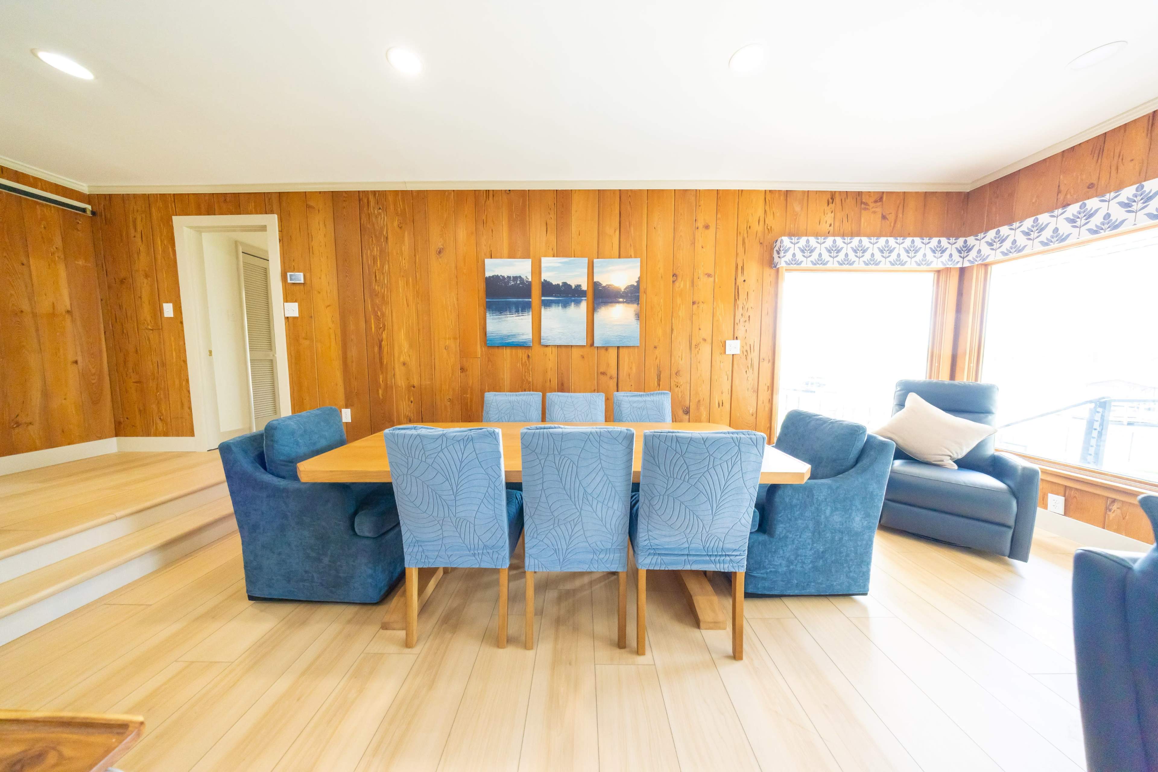 The image shows a dining area with a large wooden table surrounded by blue upholstered chairs, set against a wall of wooden panels and large windows letting in natural light.