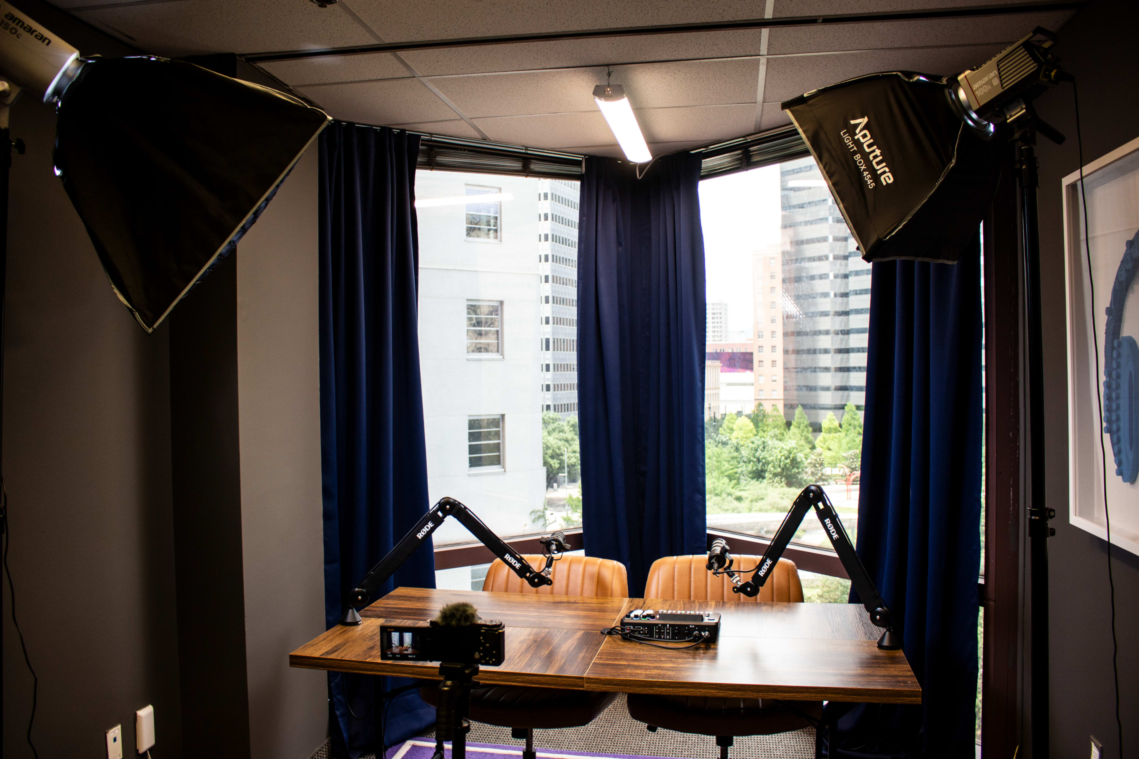 The image shows a recording studio setup with two chairs and a wooden table, flanked by professional lighting equipment and large windows overlooking a cityscape.