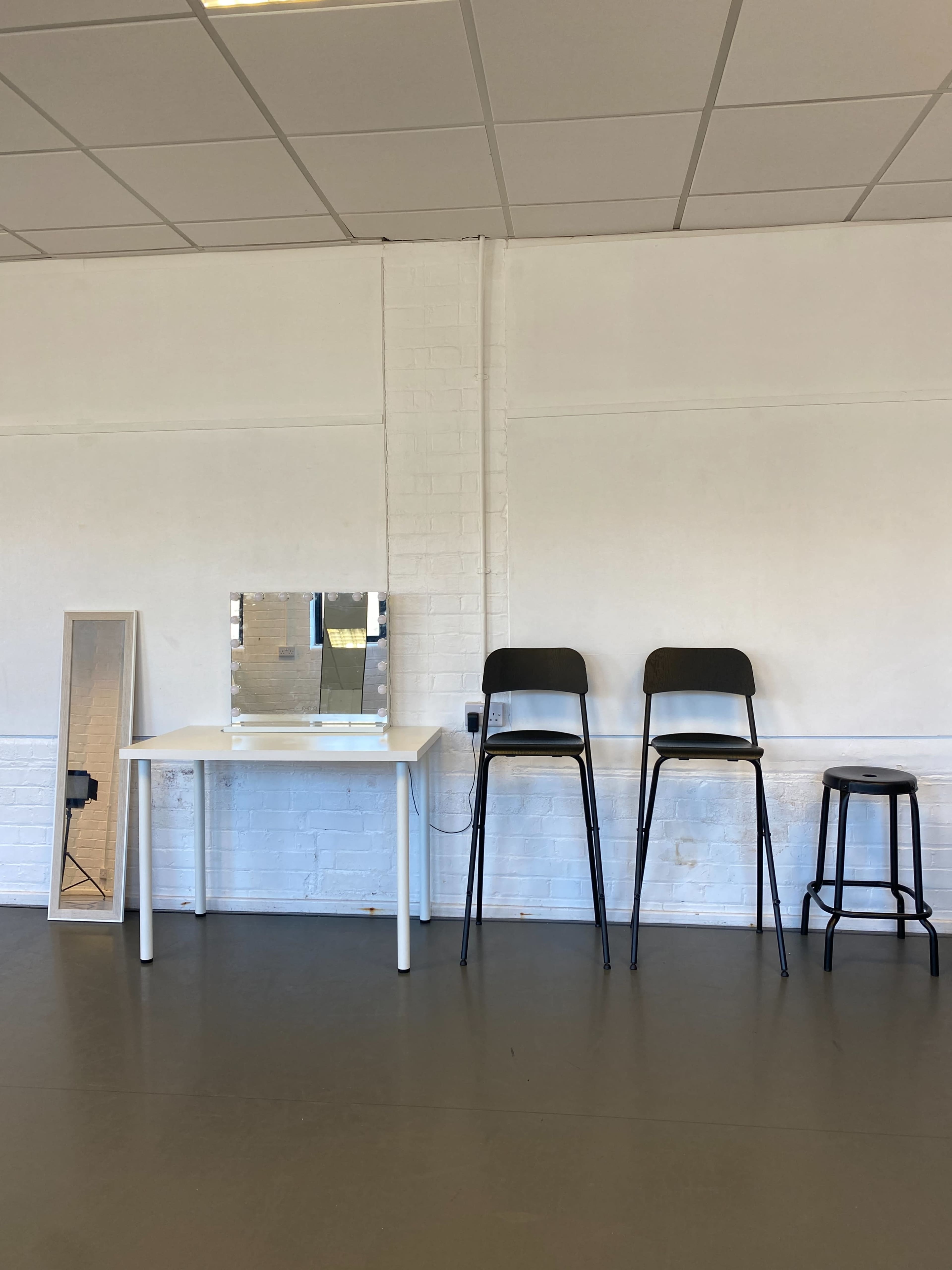 A minimalist studio space featuring a white desk with a mirror, three chairs of different designs, and a large mirror leaning against the wall.