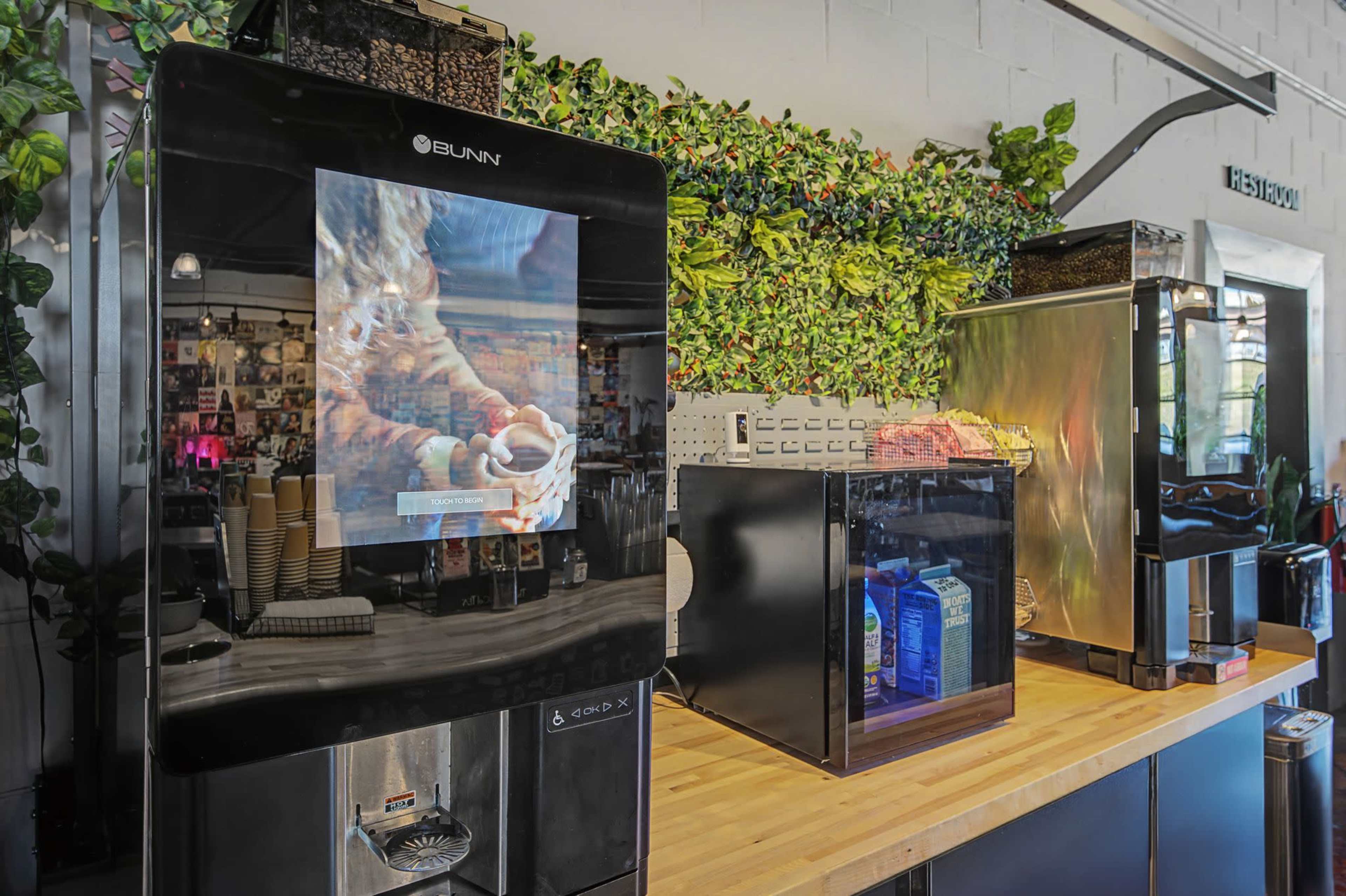 A coffee station features multiple machines, including a large BUNN coffee maker and a refrigerated unit, set against a backdrop of greenery on a wooden counter.