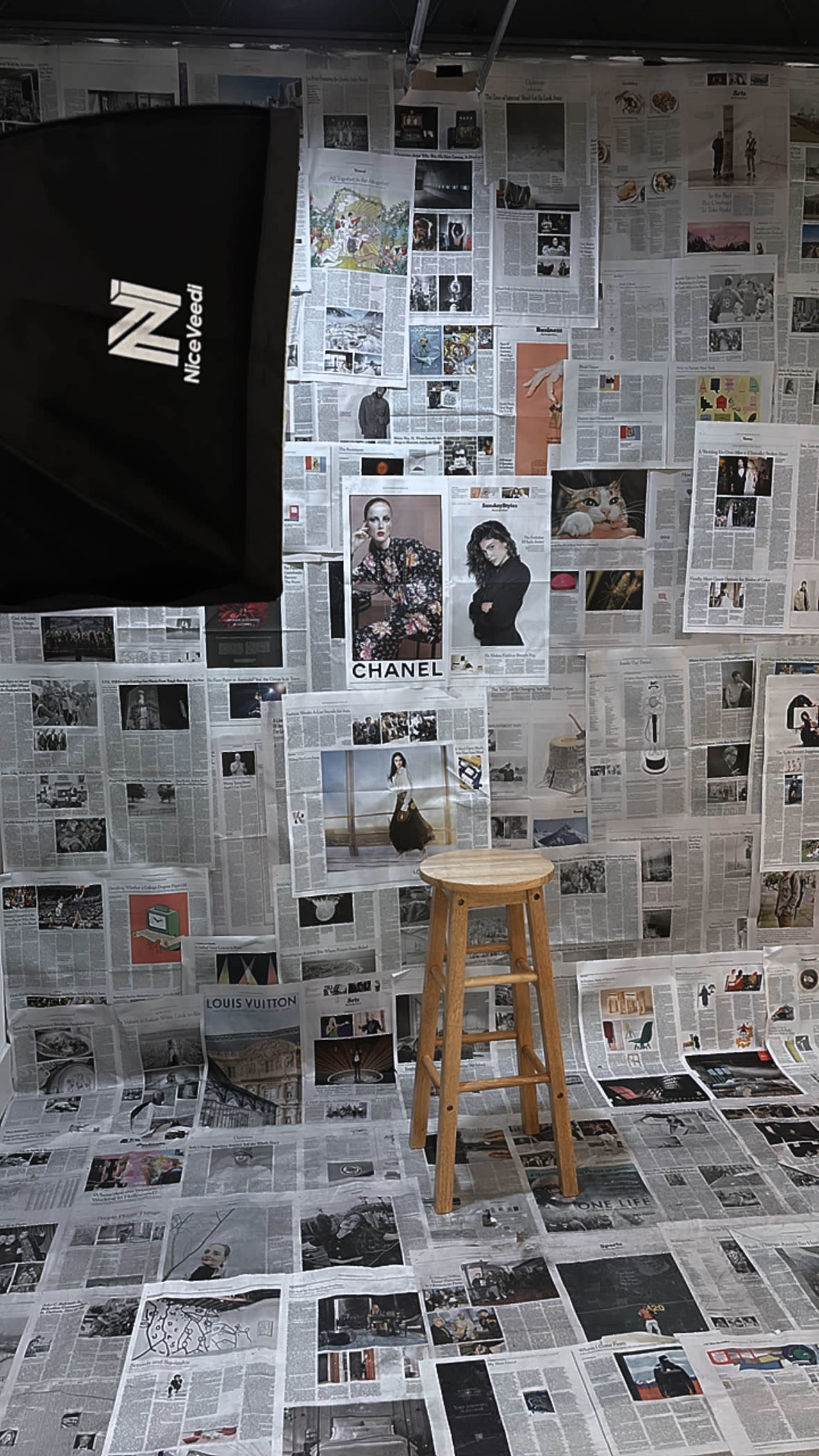 The scene features a wooden stool positioned in a room where the walls and floor are covered with newspaper clippings and photographs, including images of Chanel and Louis Vuitton.