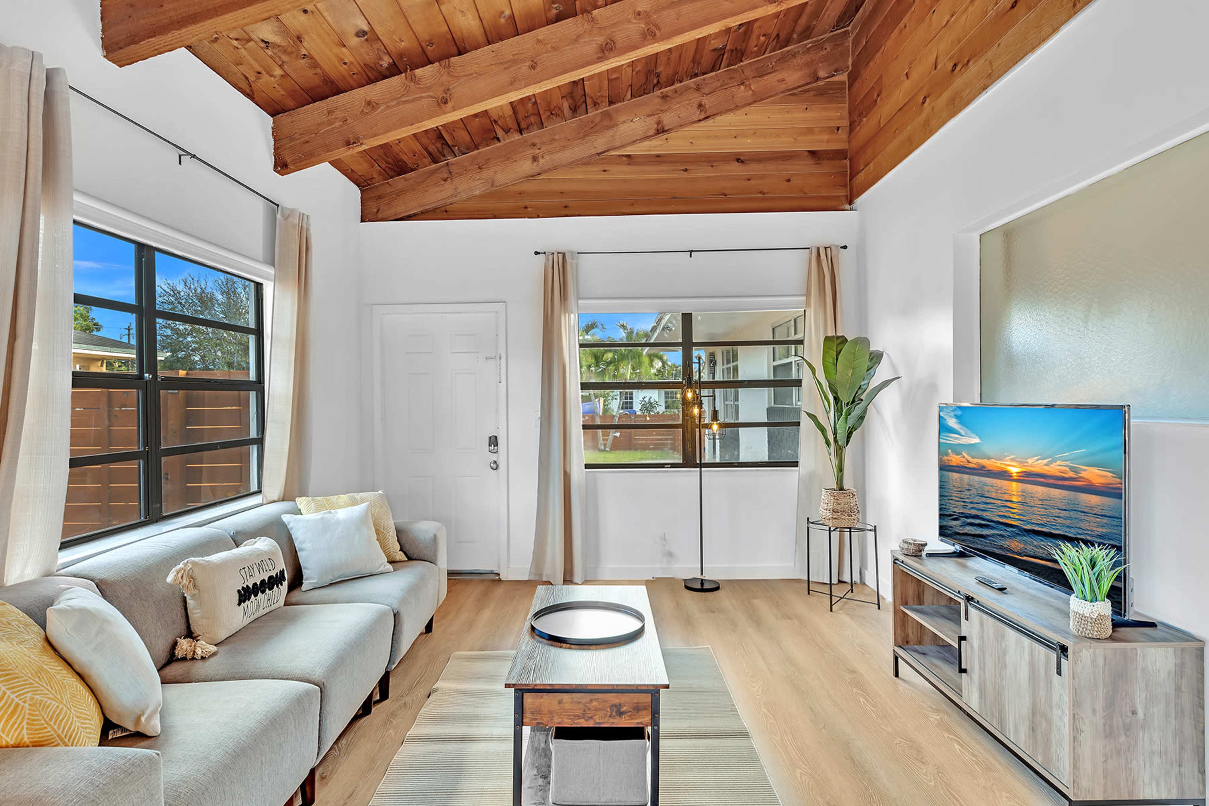 A living room features a sofa, a coffee table, a TV stand with a television, large windows, and a door leading outside, all under a wooden-beamed ceiling.