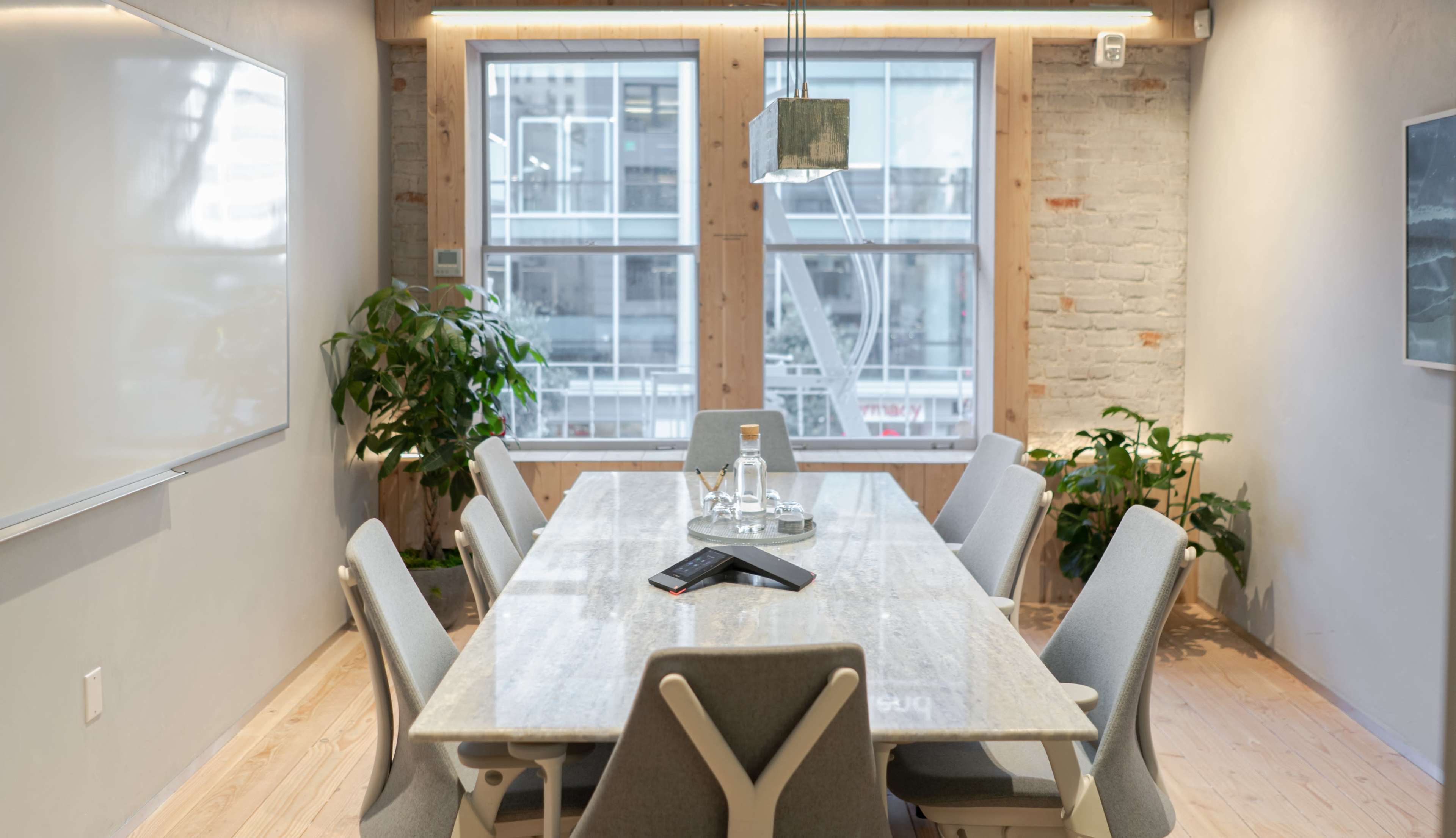 A modern conference room features a long table surrounded by chairs, with large windows allowing natural light and plants positioned along the walls.