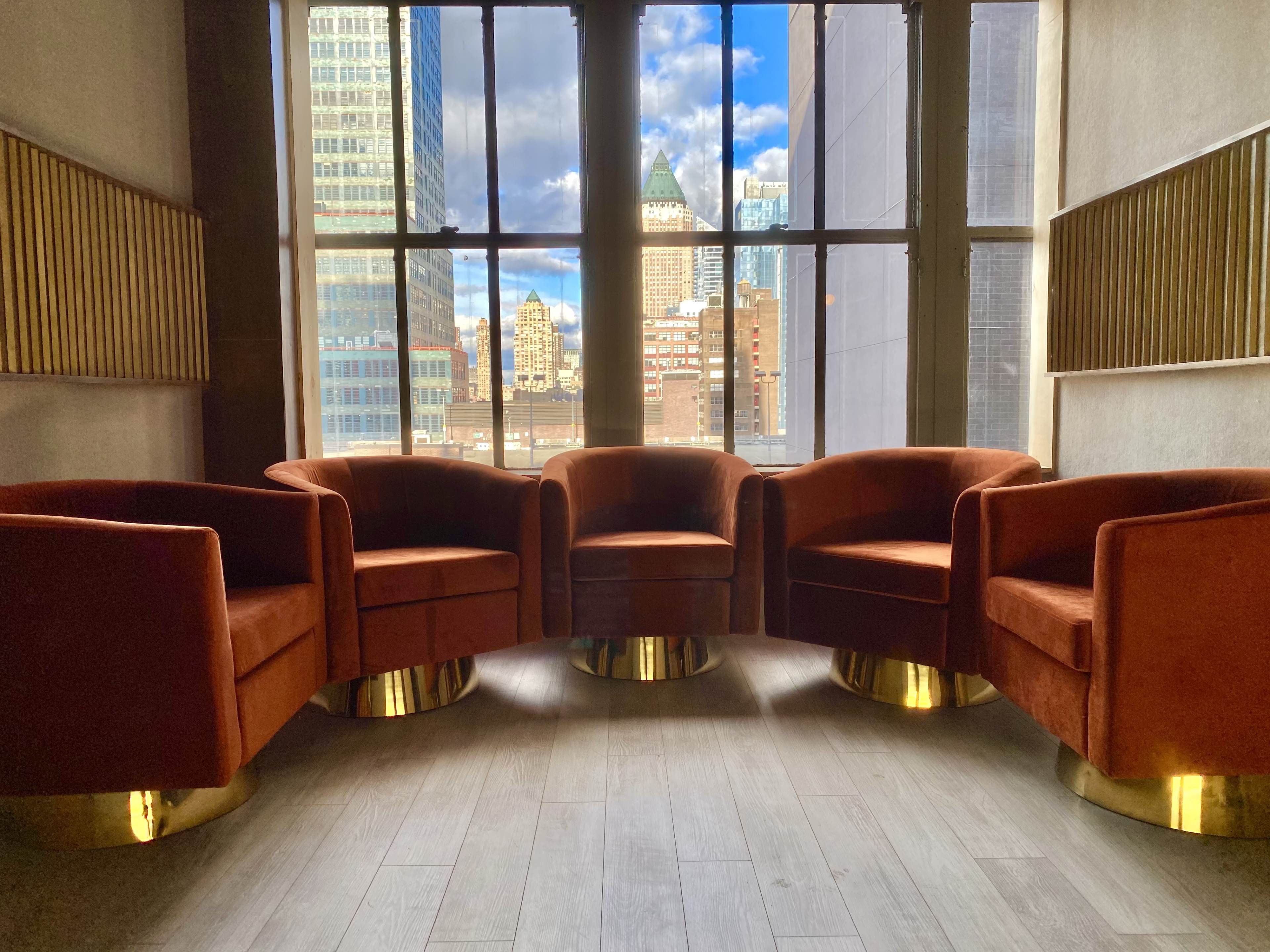 Four orange velvet chairs with gold bases are arranged in a circular formation near large windows that offer a view of a city skyline.