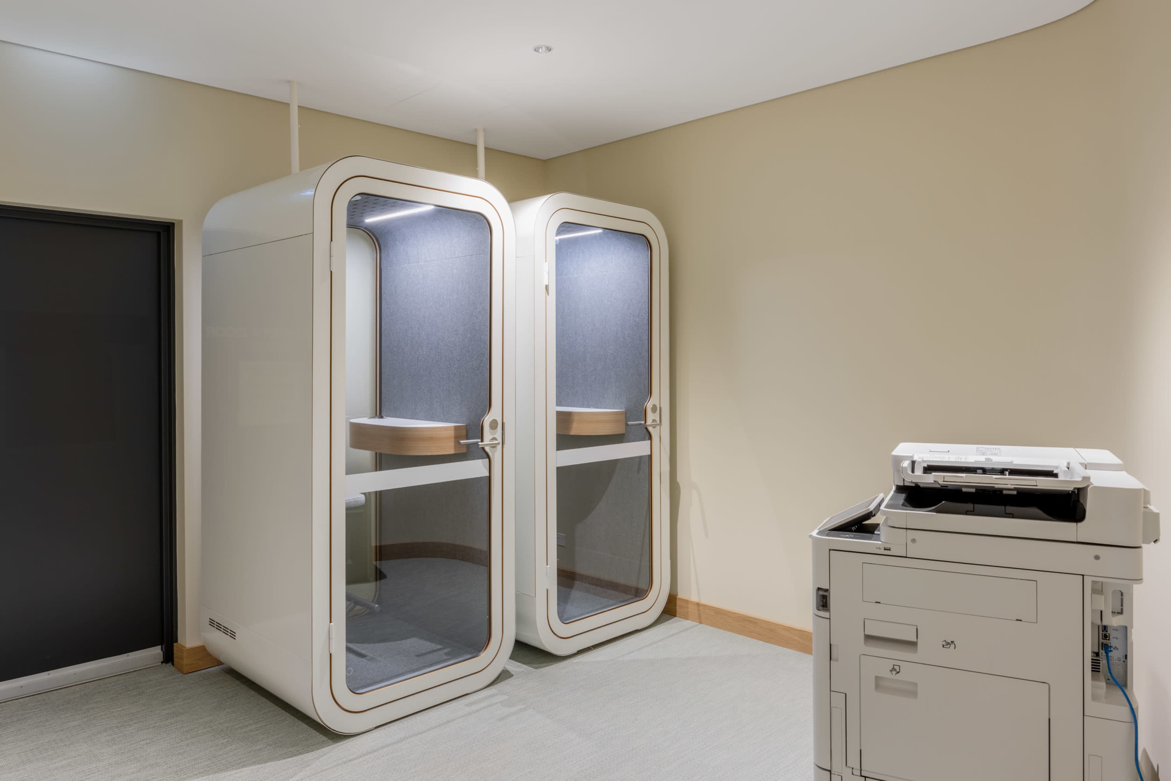 The image shows two soundproof phone booths set against a plain wall with a nearby multifunction printer.