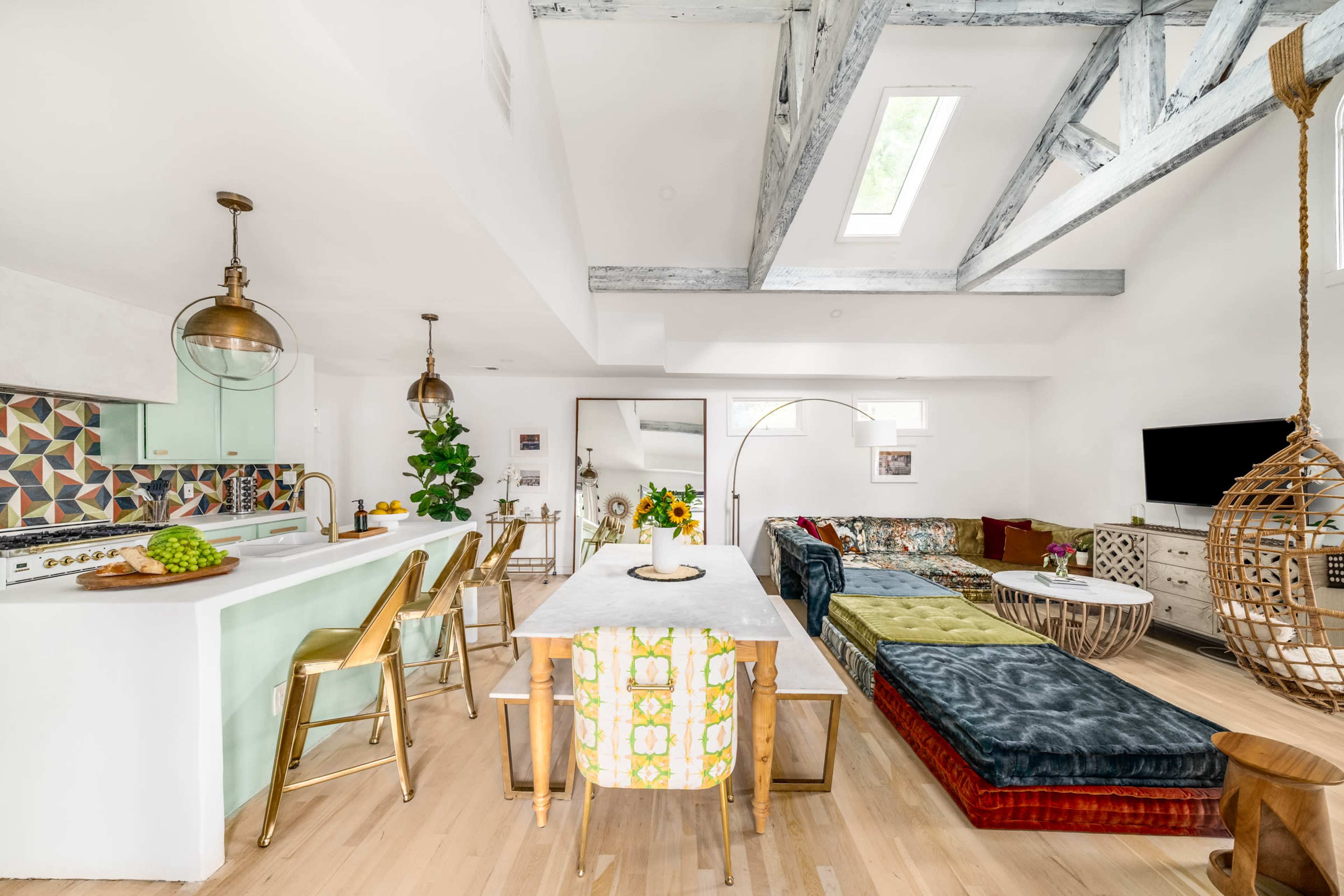 A modern living space features a kitchen with a bar, a dining table, and a colorful seating area, all under exposed wooden beams and large windows.