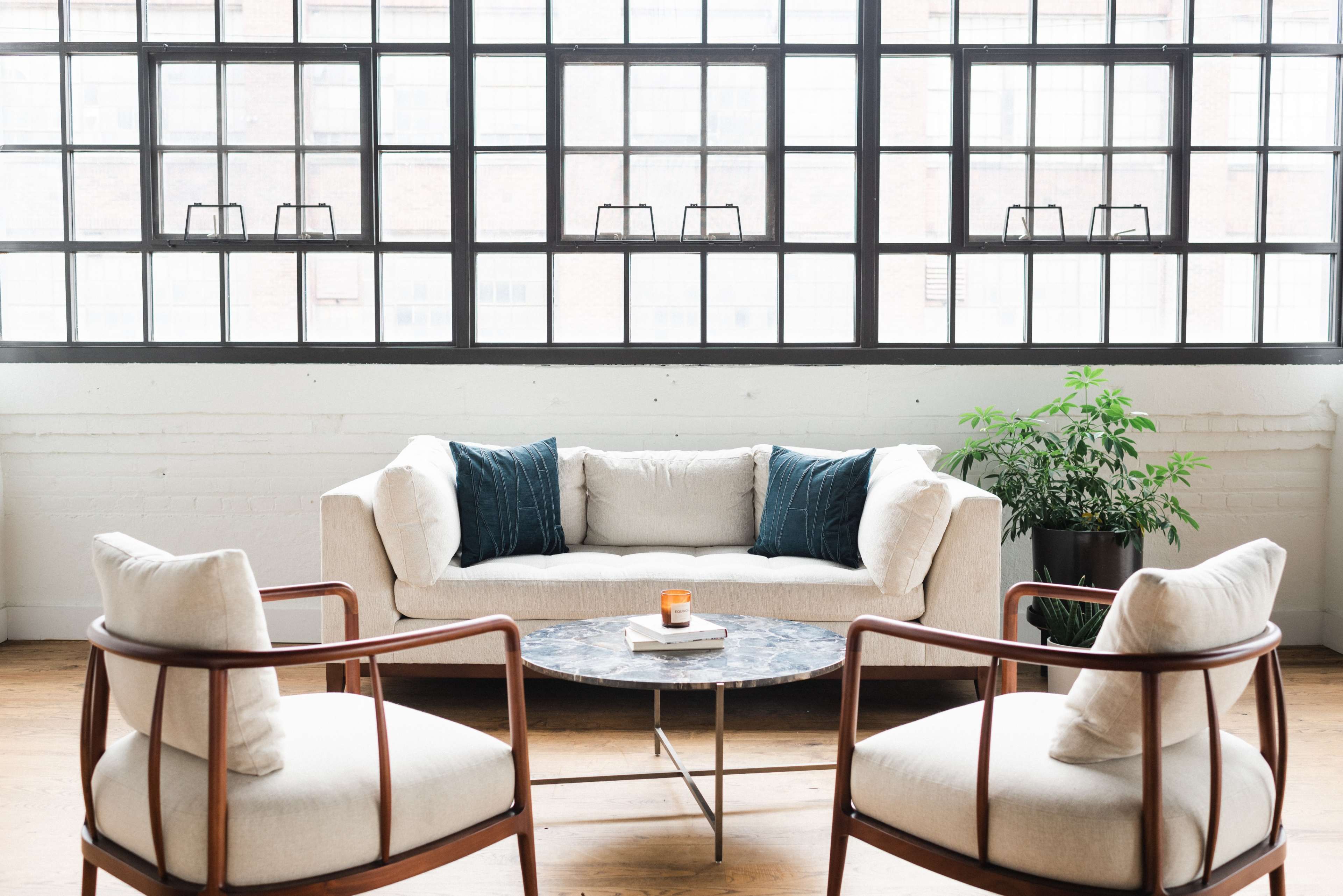 A light-colored sectional sofa paired with two wooden chairs and a round marble coffee table sits in front of large industrial windows.