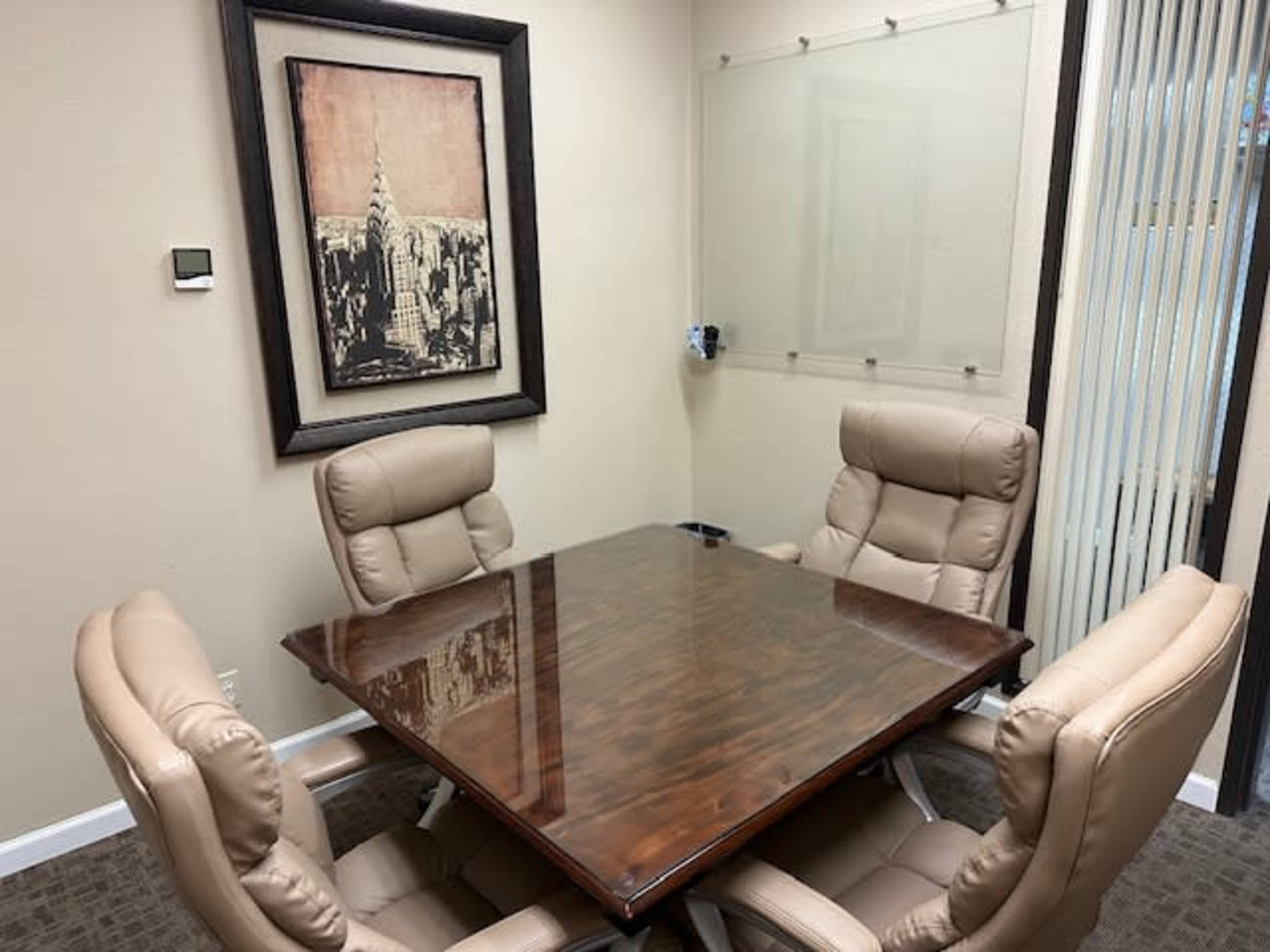 A small conference room features a wooden table surrounded by four beige swivel chairs and a wall-mounted artwork.