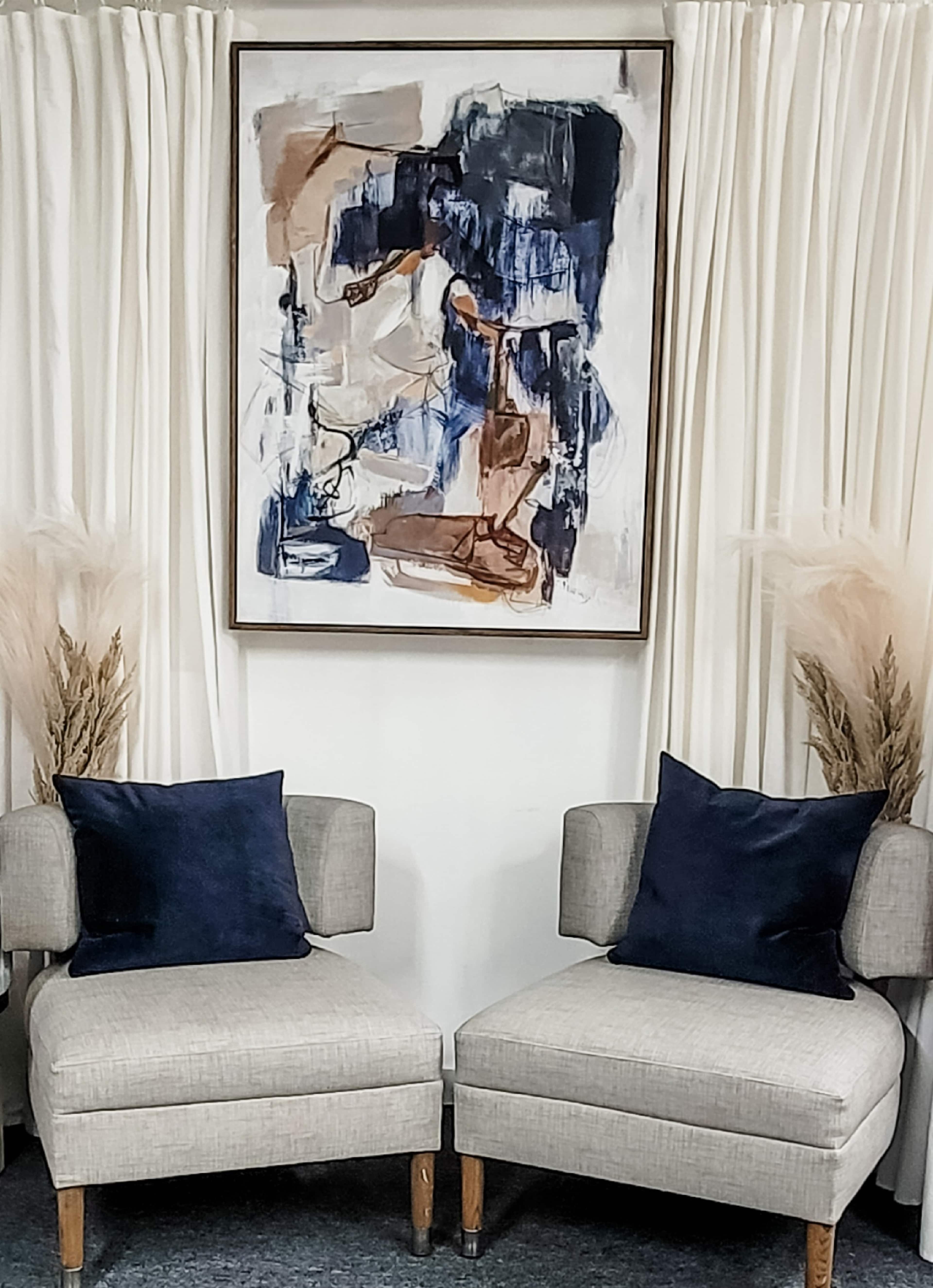 Two light gray chairs with dark blue cushions are positioned facing each other beneath a large abstract painting, flanked by white curtains and decorative dried plants.