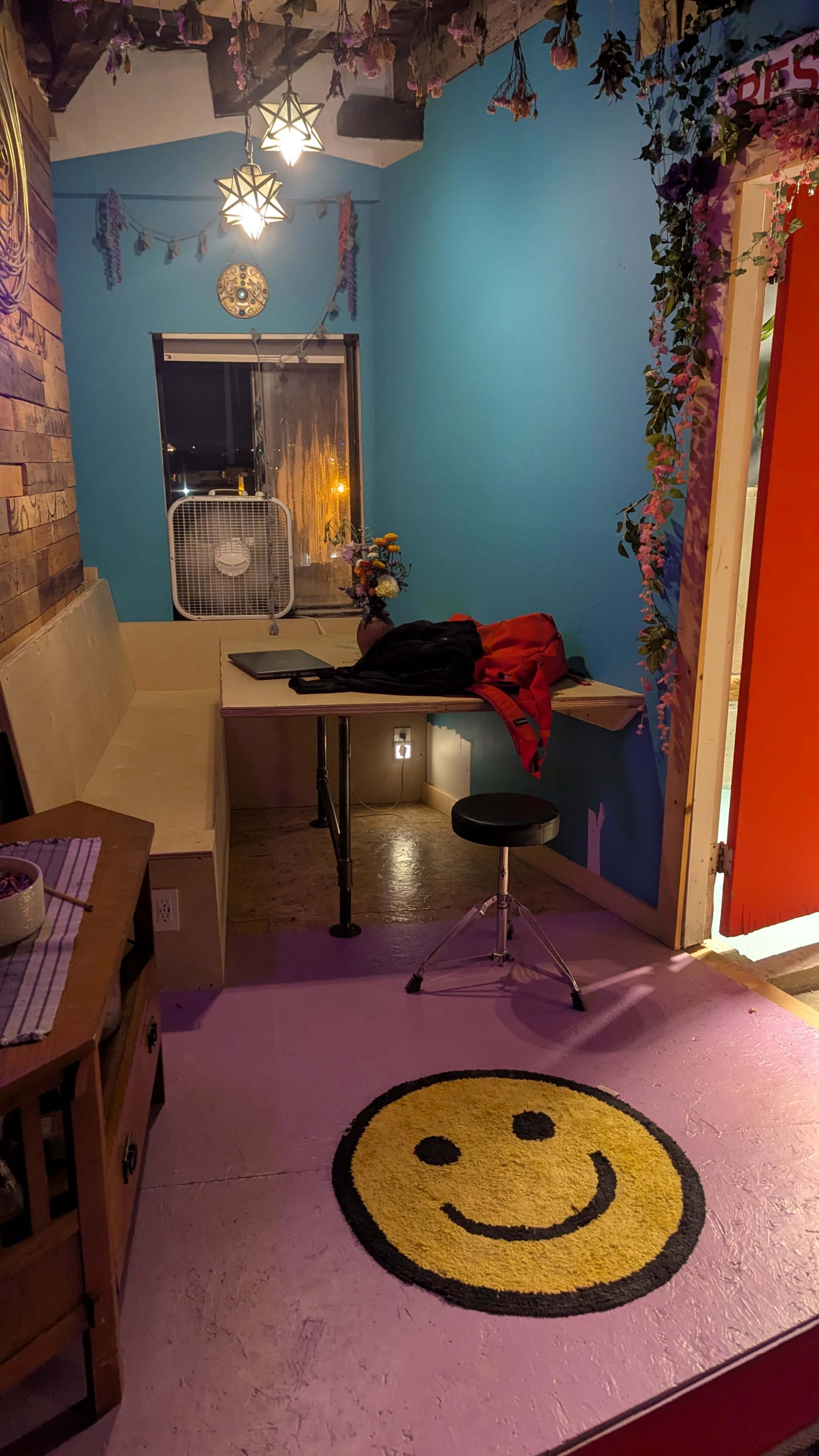 The image shows a small, colorful room with a wooden table, a chair, a fan, and a bright red door.