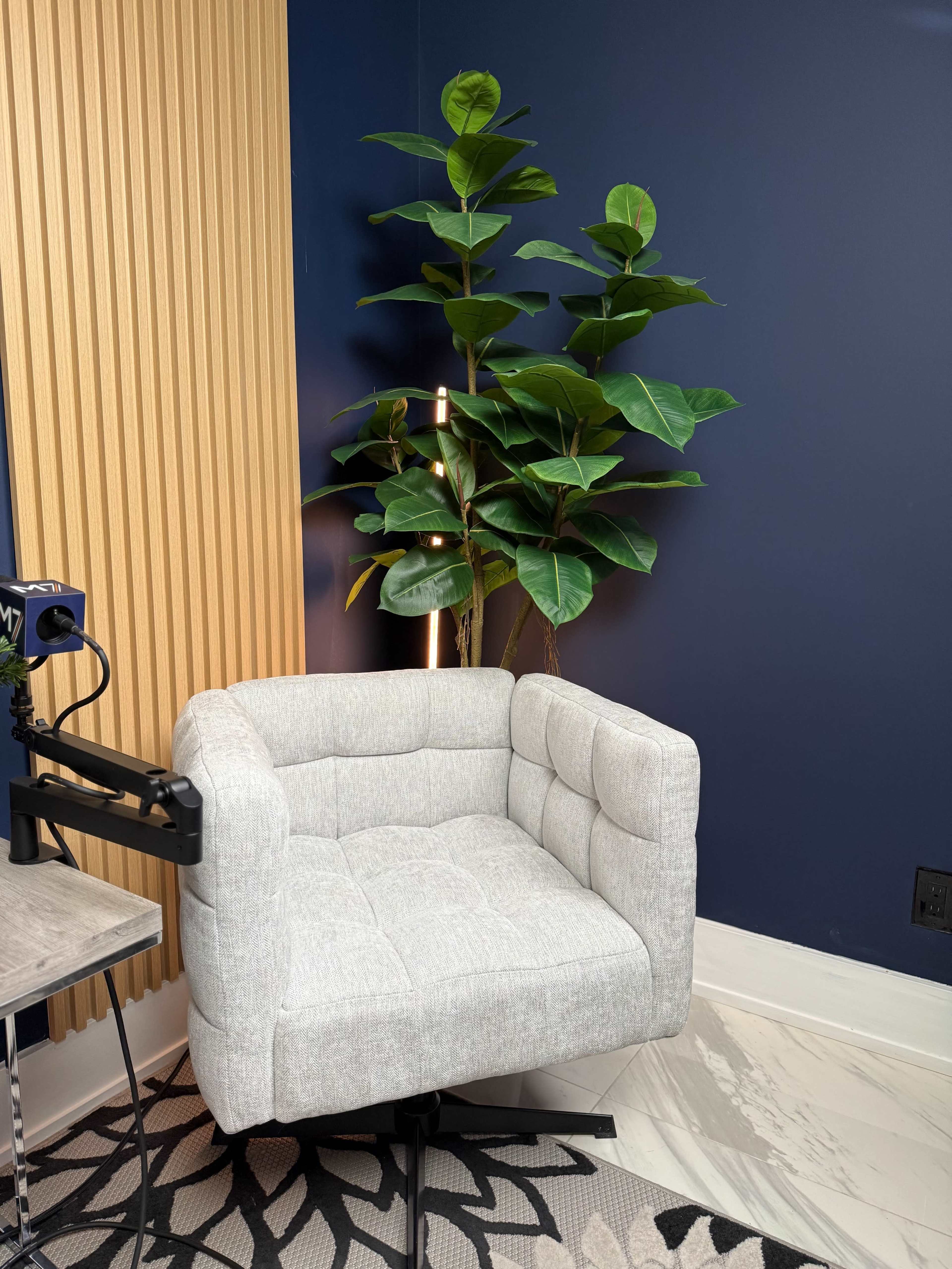 A light gray, tufted armchair is positioned beside a tall, green plant against a dark blue wall, with a wooden accent panel in the background.
