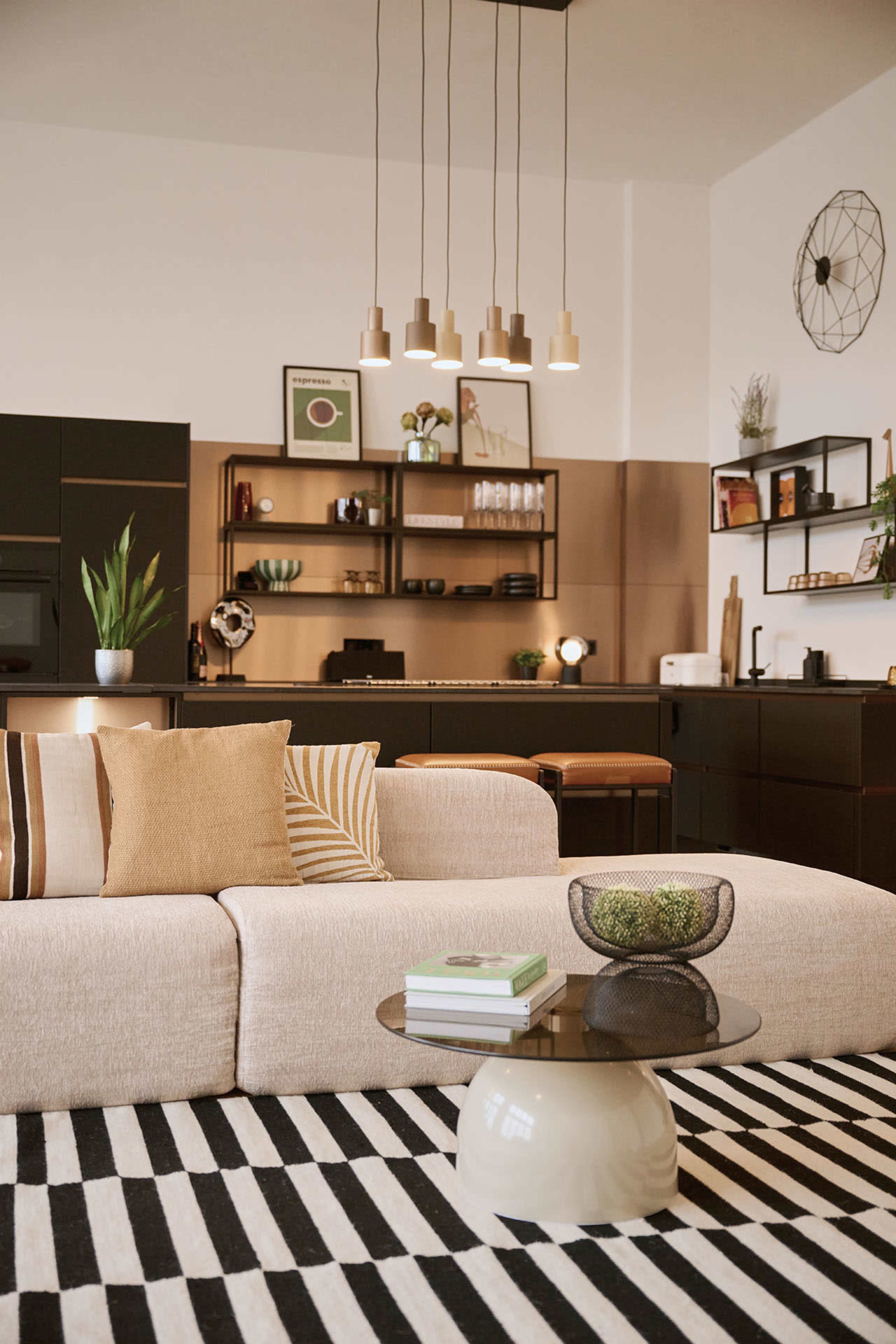 The image shows a modern living room with a beige sofa, a striped black and white rug, and a contemporary coffee table, with a kitchen area visible in the background featuring pendant lighting and plants.