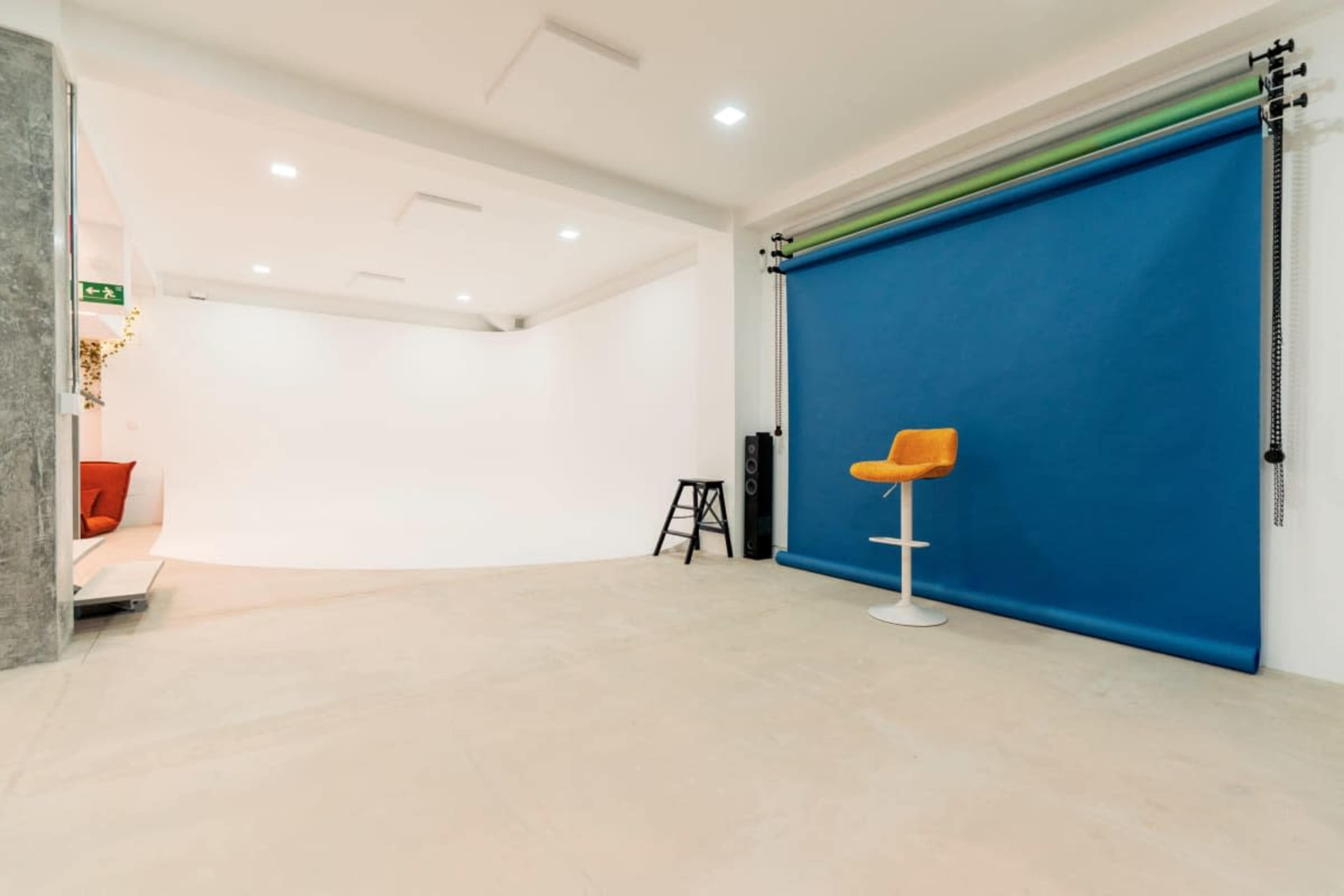 The image shows a spacious photography studio with a blue backdrop, a single orange chair, and a gray wall.