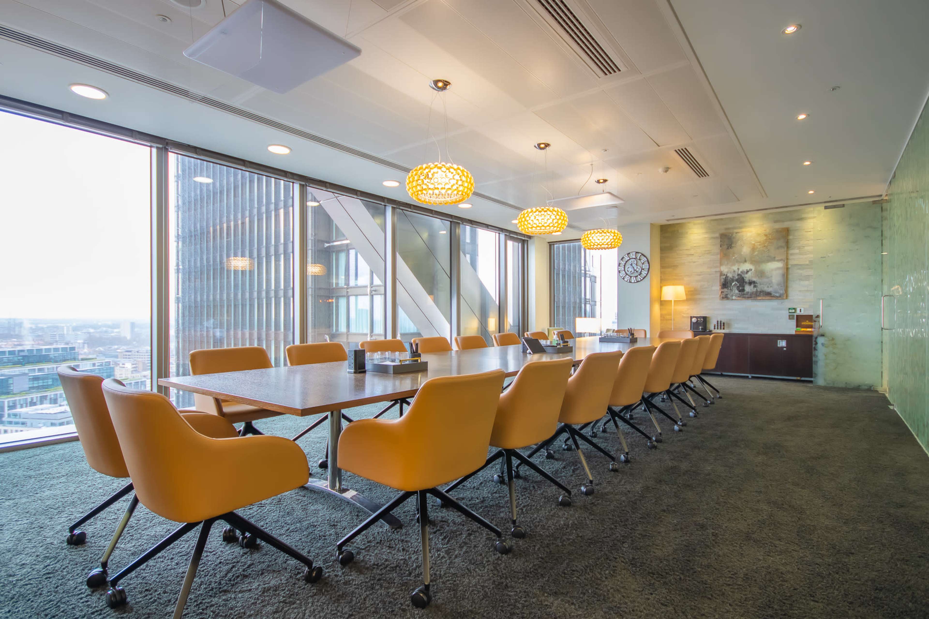 A modern conference room with a large wooden table surrounded by orange chairs, large windows, and decorative lighting fixtures.