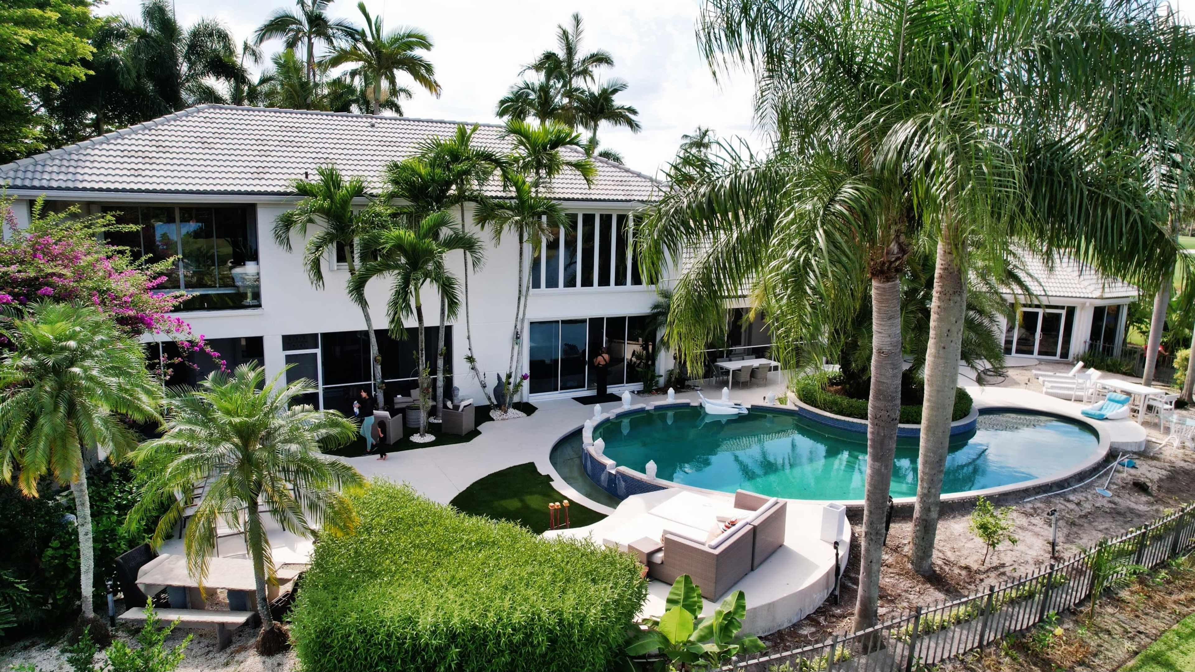 A modern two-story house surrounded by palm trees, a swimming pool, and landscaped gardens.