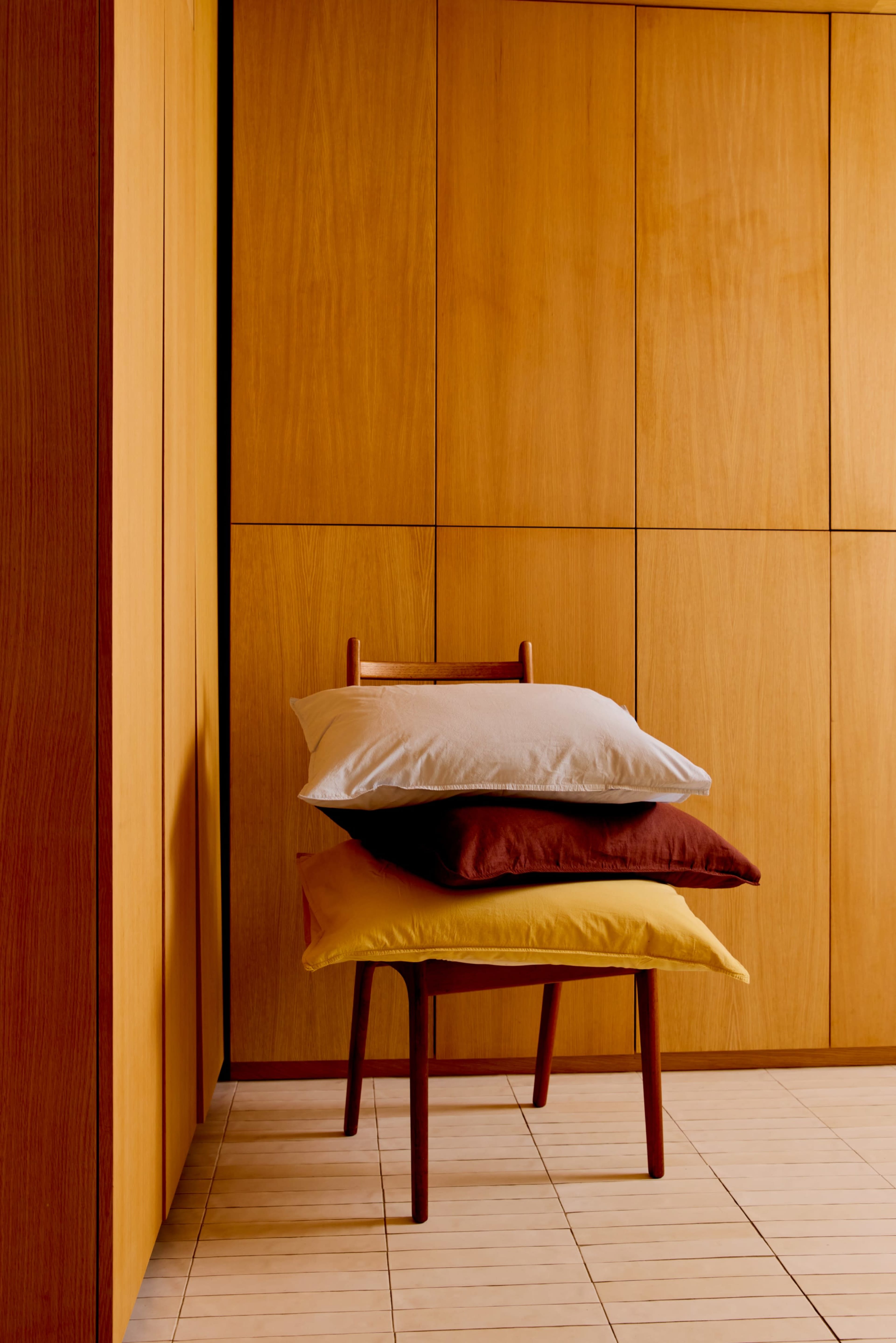 A wooden chair with three stacked pillows in varying colors is positioned against a backdrop of wooden wall panels.