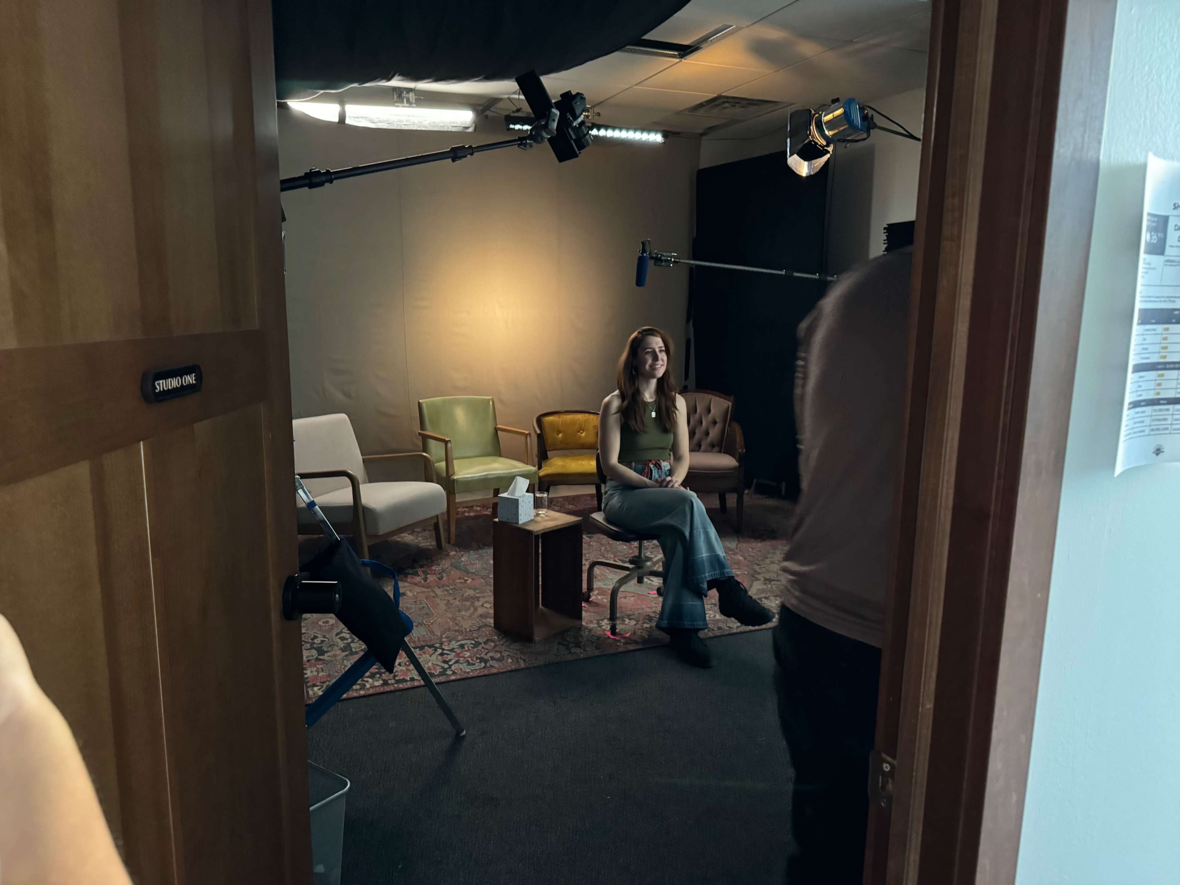 A woman sits in a chair in a studio with various chairs and lighting equipment set up around her.