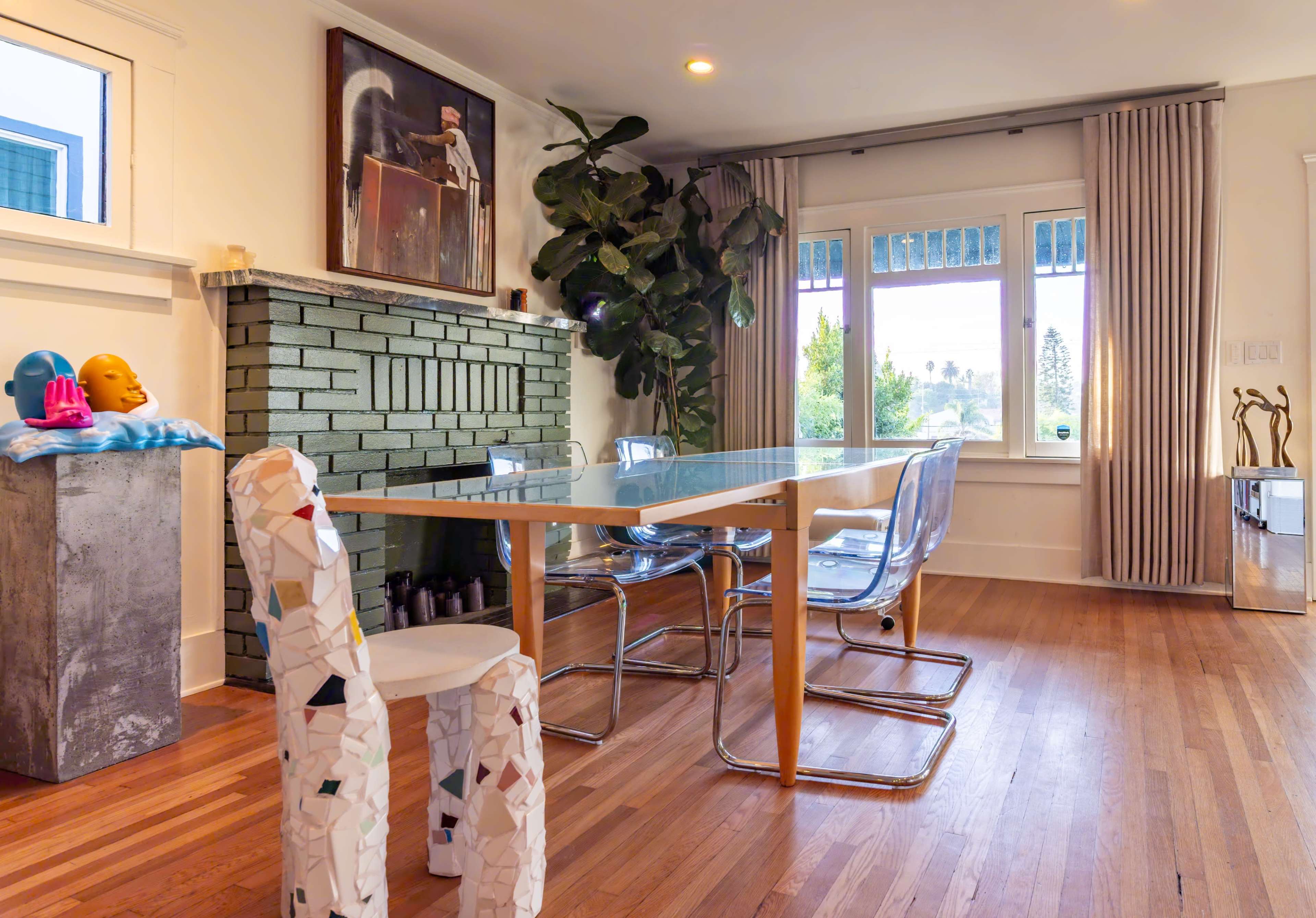 The image shows a modern dining area with a glass table surrounded by transparent chairs, a minimalist stone fireplace, and large windows letting in natural light.