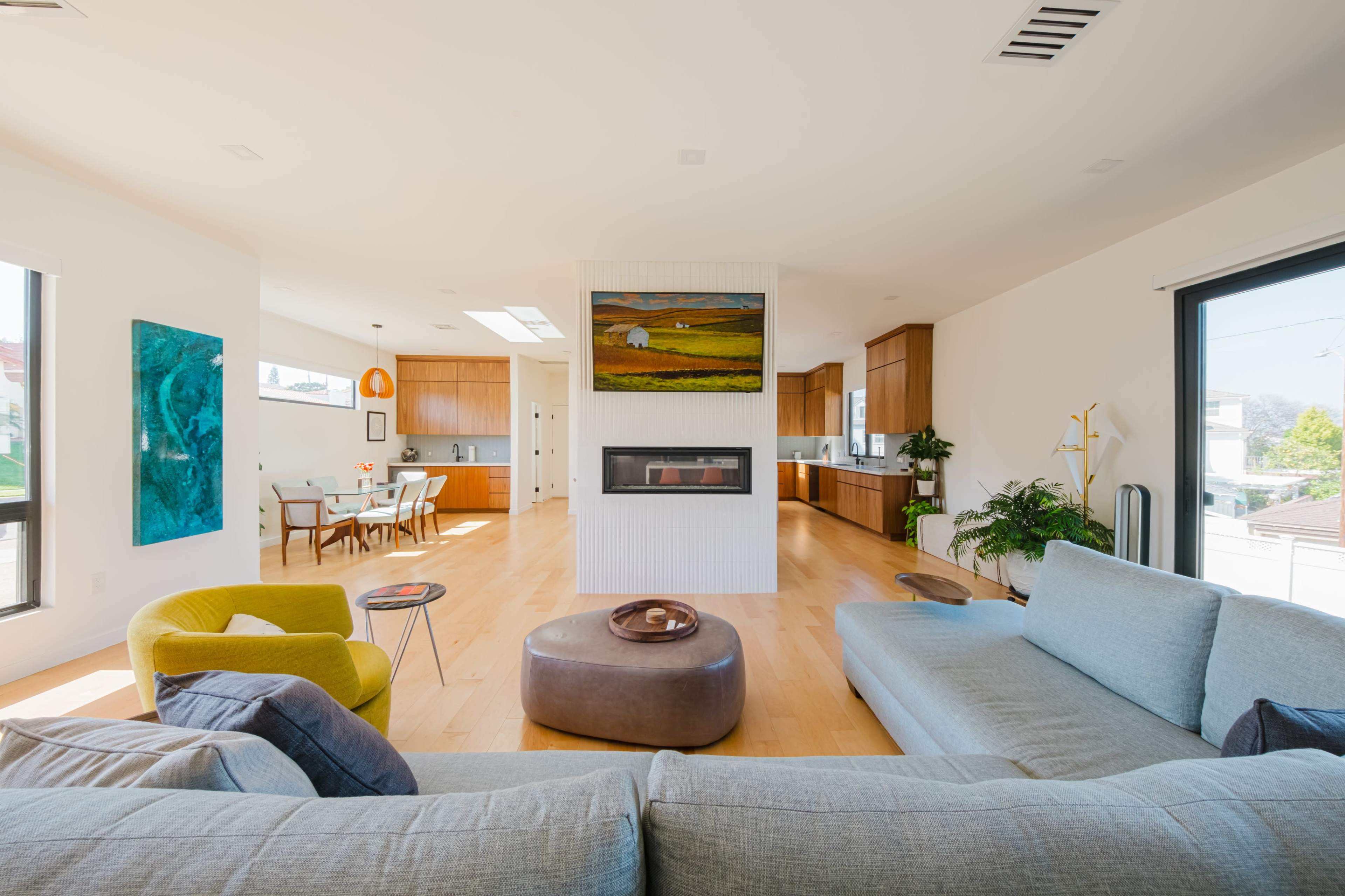 The image shows a spacious, modern living room with large windows, contemporary furniture, and an open layout that connects to a dining area and kitchen.