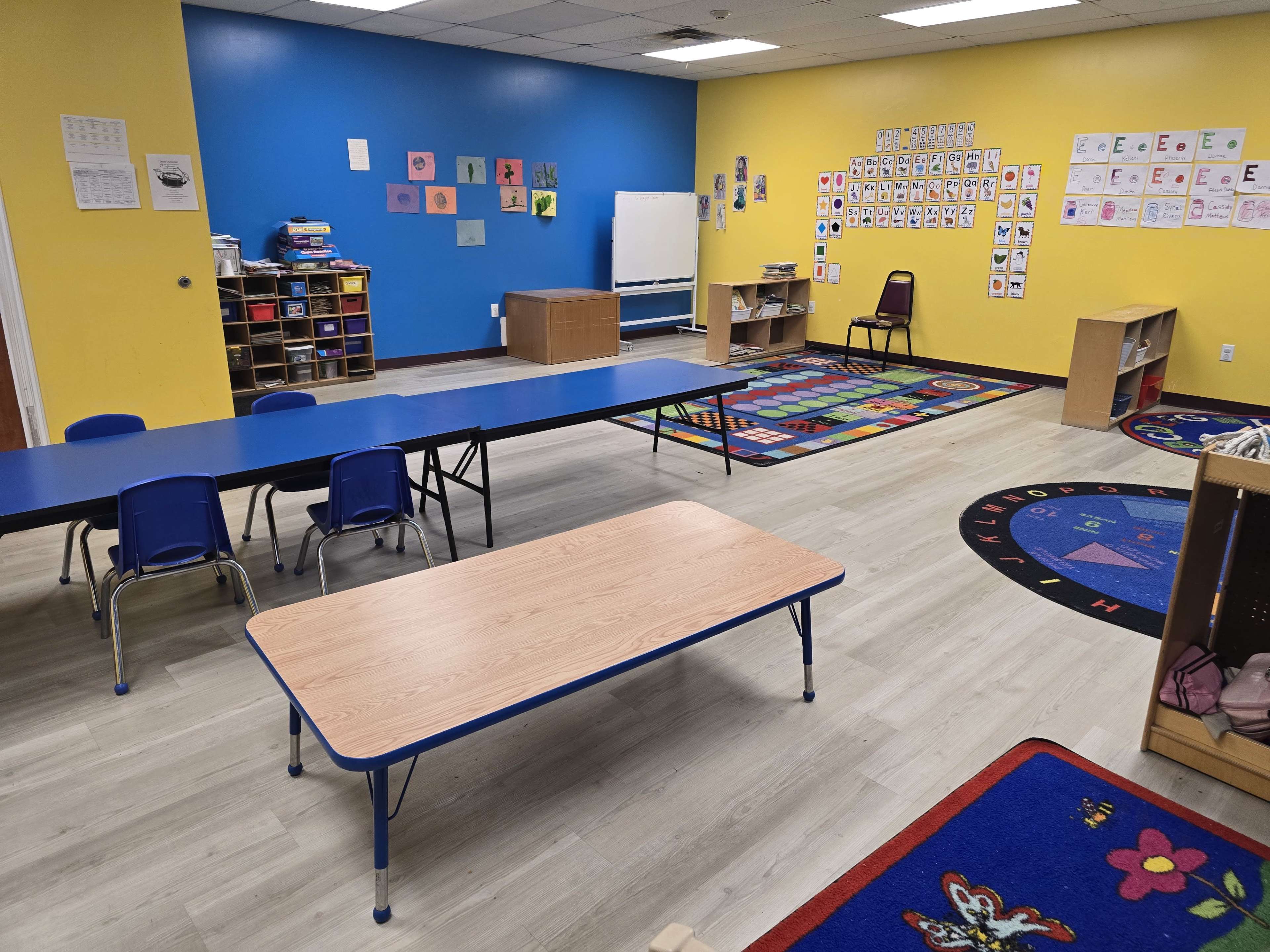 The image shows a colorful classroom with blue and yellow walls, including tables, chairs, a rug, and educational materials displayed on the walls.