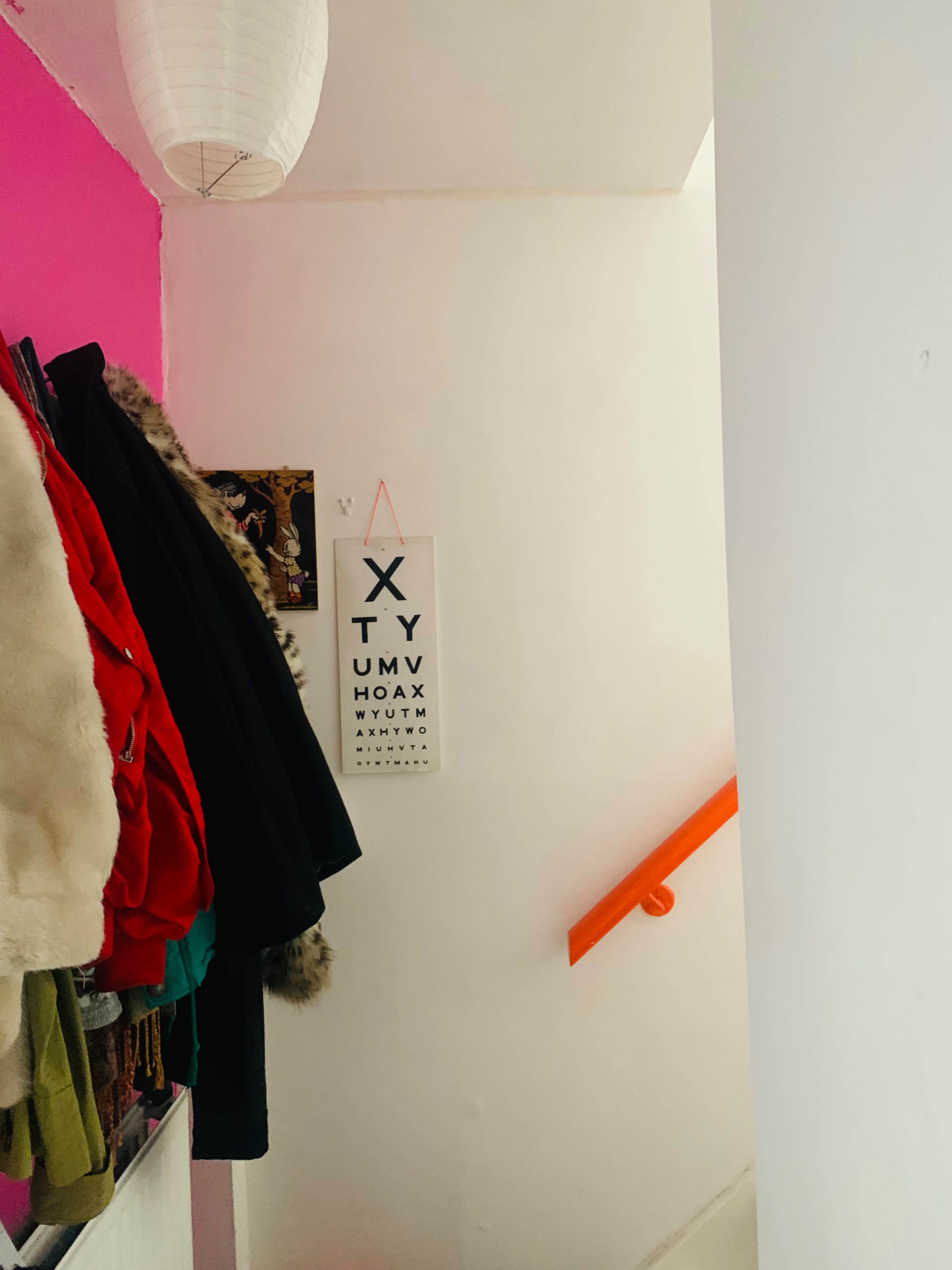 A narrow staircase with a colorful wall, a hanging eye chart, and a row of coats on the left.