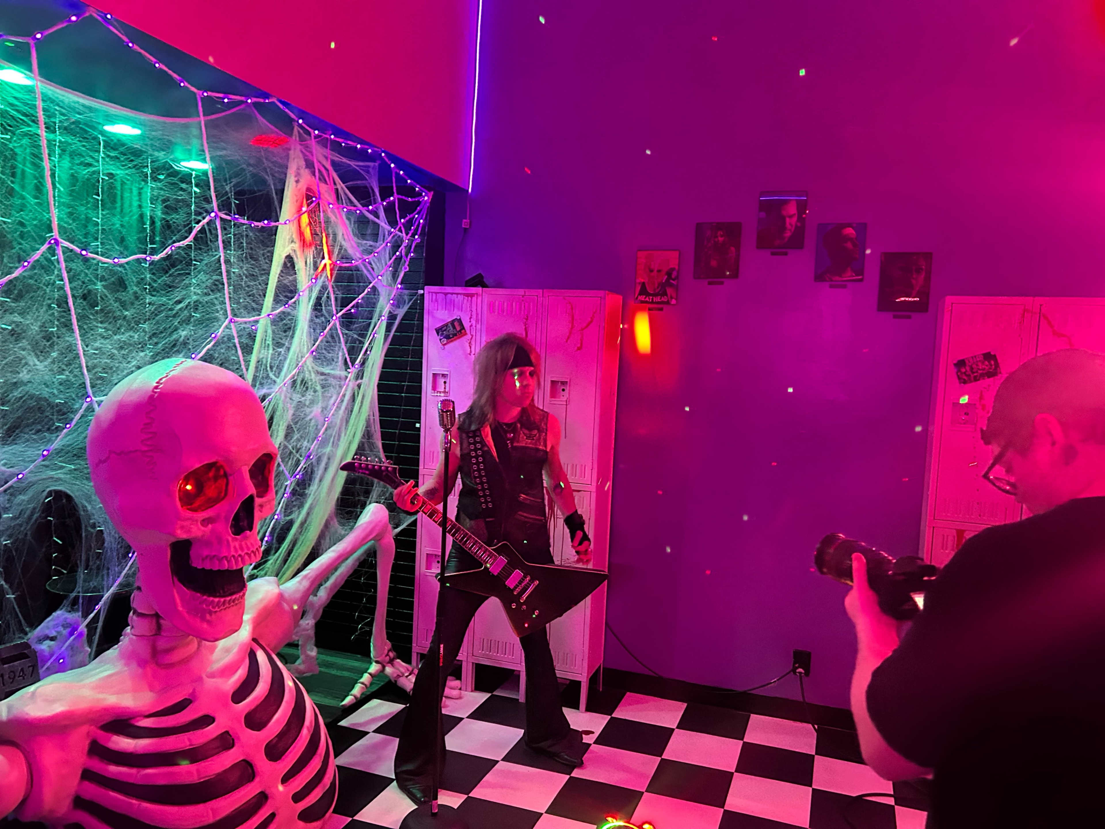 A person dressed in a rock outfit poses with a guitar alongside a skeleton in a brightly lit, colorful room decorated for Halloween.