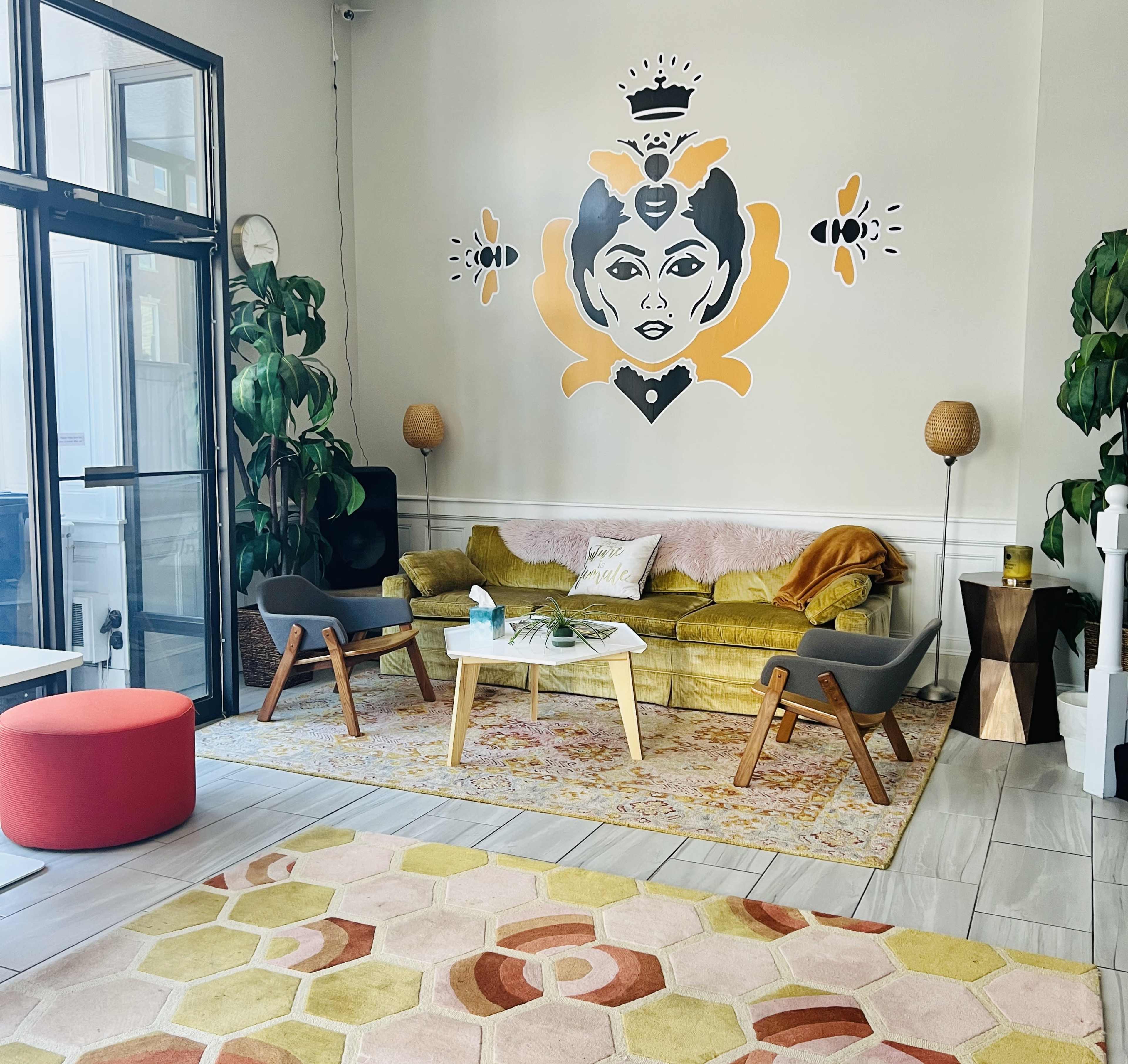 The scene features a modern living space with a yellow sofa, decorative wall art, chairs, a coffee table, and plants, all arranged on a patterned rug.