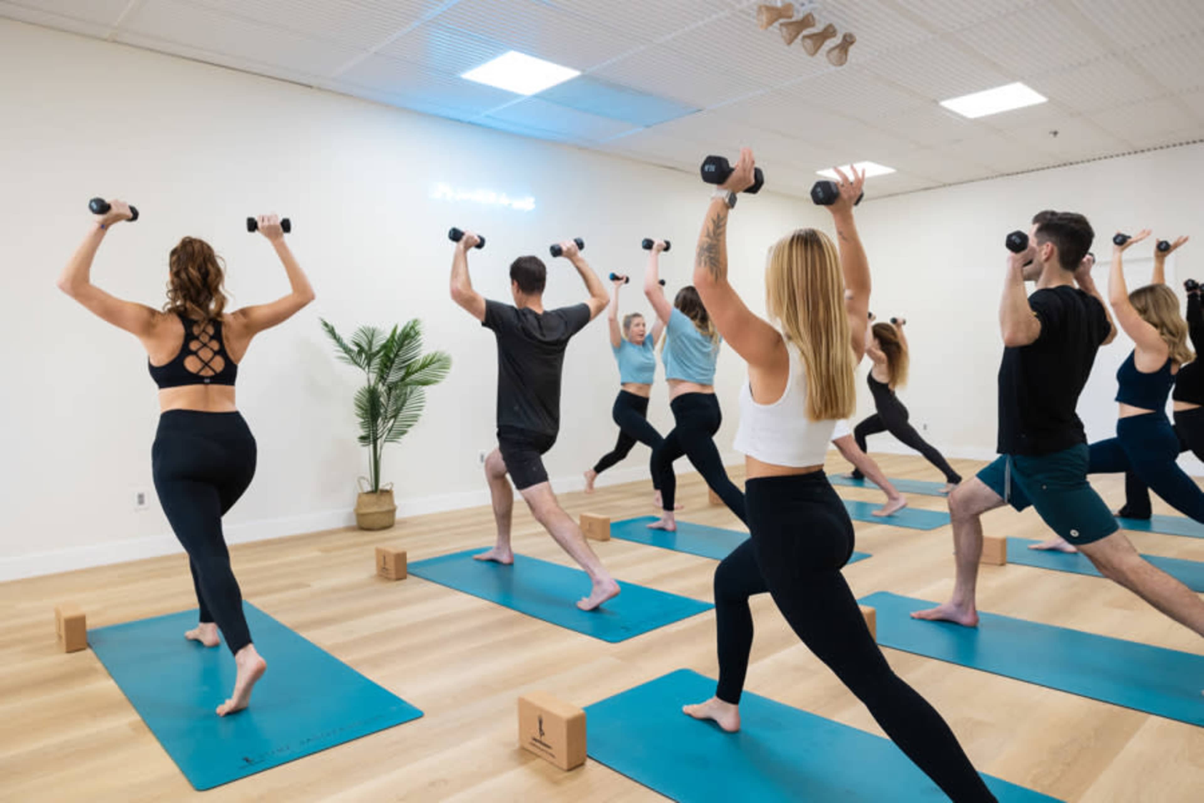 A group of people is participating in a yoga class, using weights while positioned on yoga mats in a bright studio.