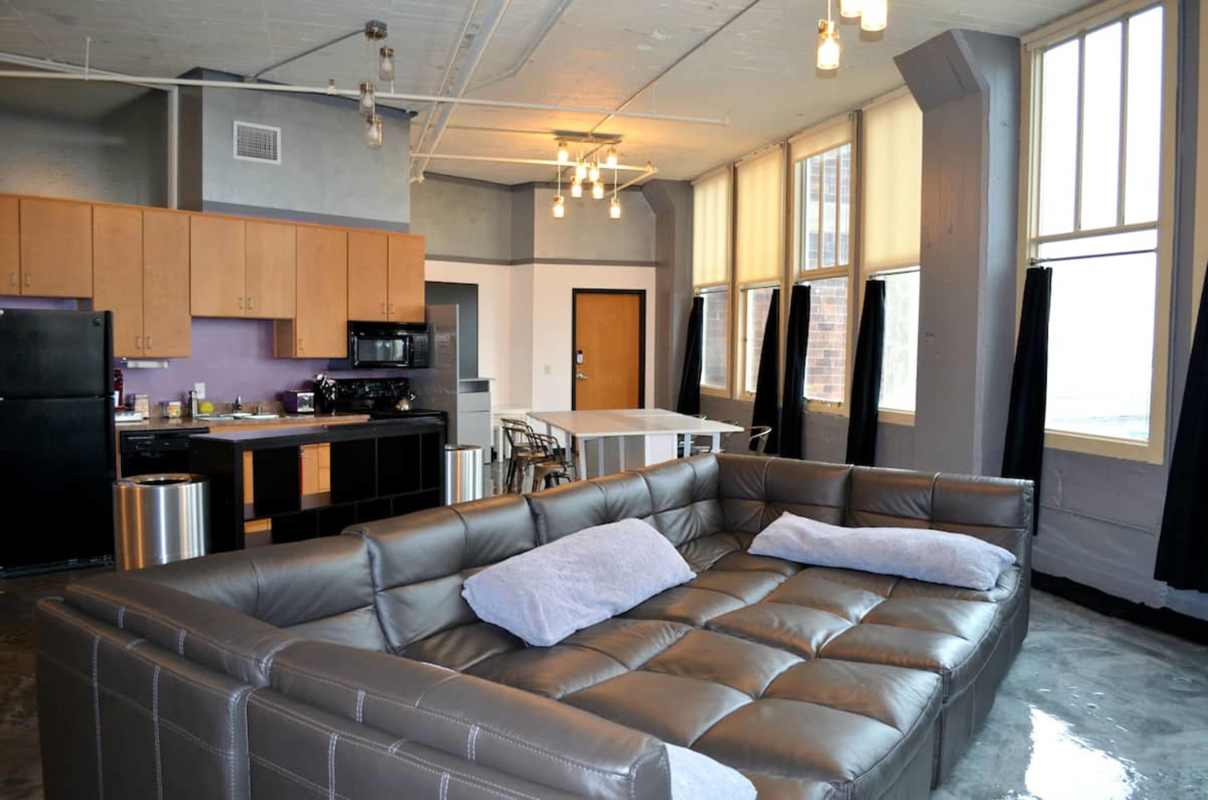 A modern living space featuring a large sectional sofa, a kitchen with wooden cabinets, and a dining area with windows.