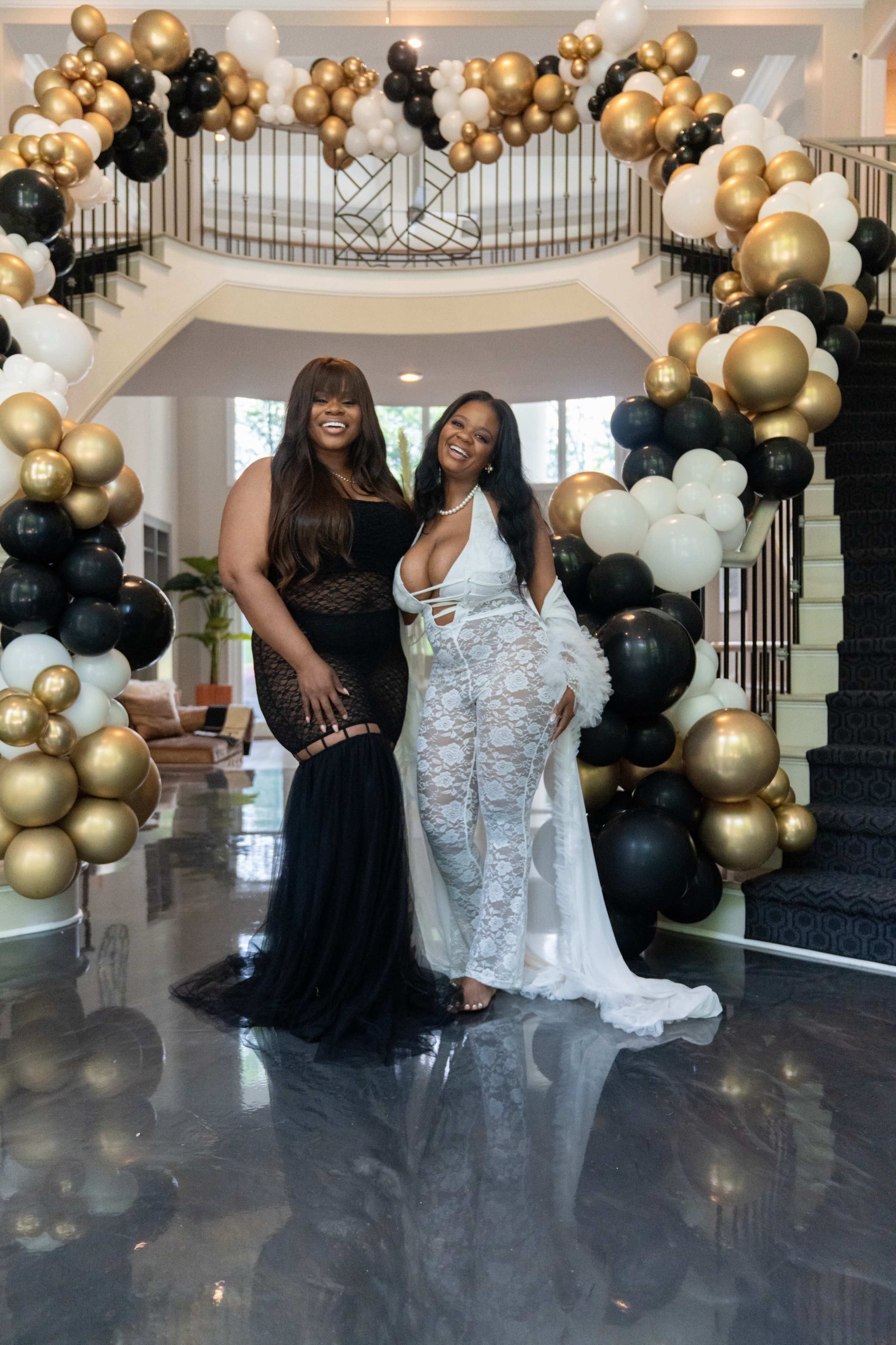 Two women pose together in an elegantly decorated entryway with a large arch made of black, white, and gold balloons.