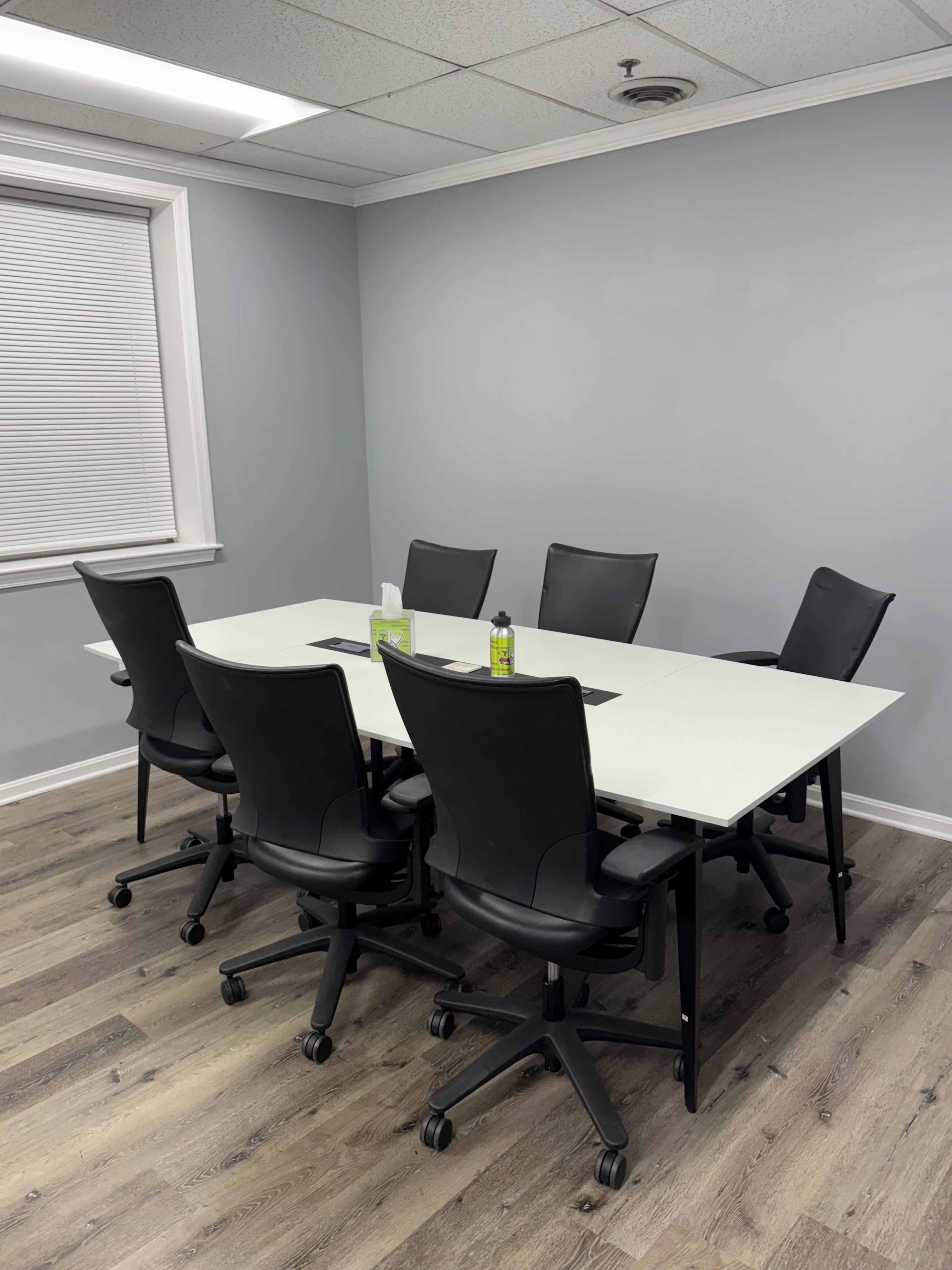 A conference room features a rectangular white table surrounded by six black office chairs.