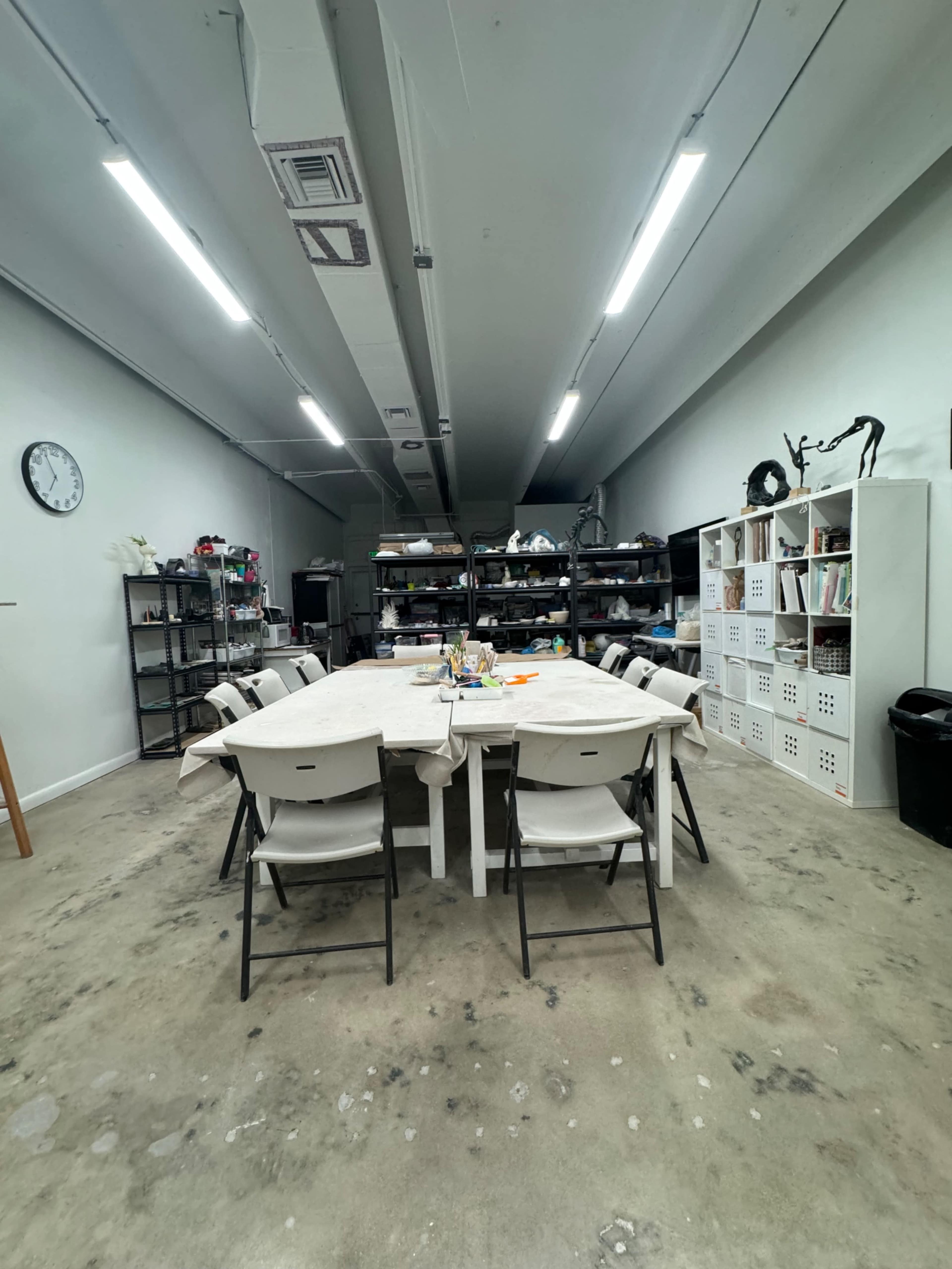 A long table with chairs sits in a spacious, well-lit room that features shelving units and a clock on the wall.
