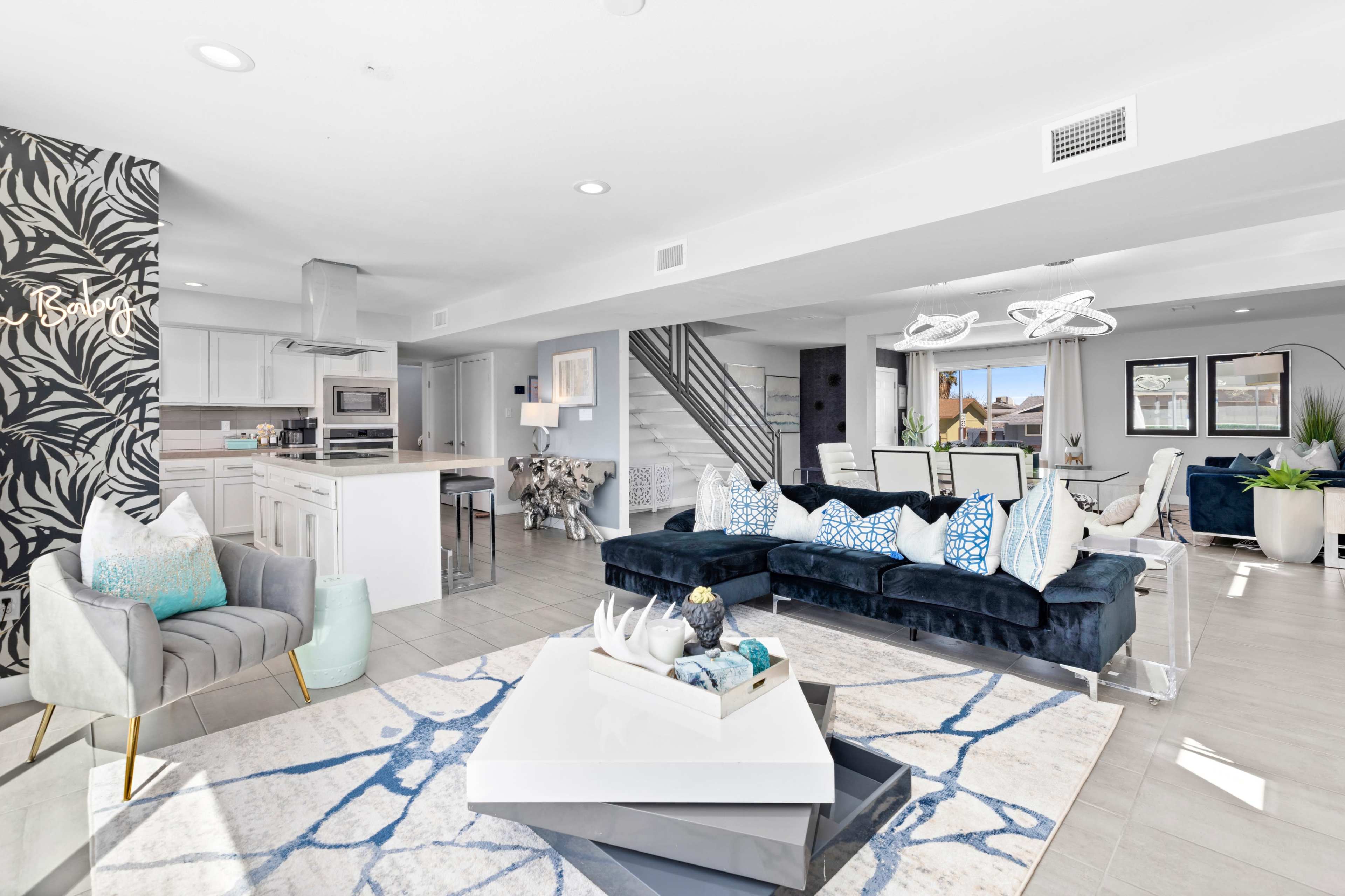 A modern open-concept living space features a dark blue sectional sofa, a coffee table with decorative items, and a stylish accent wall with tropical foliage.