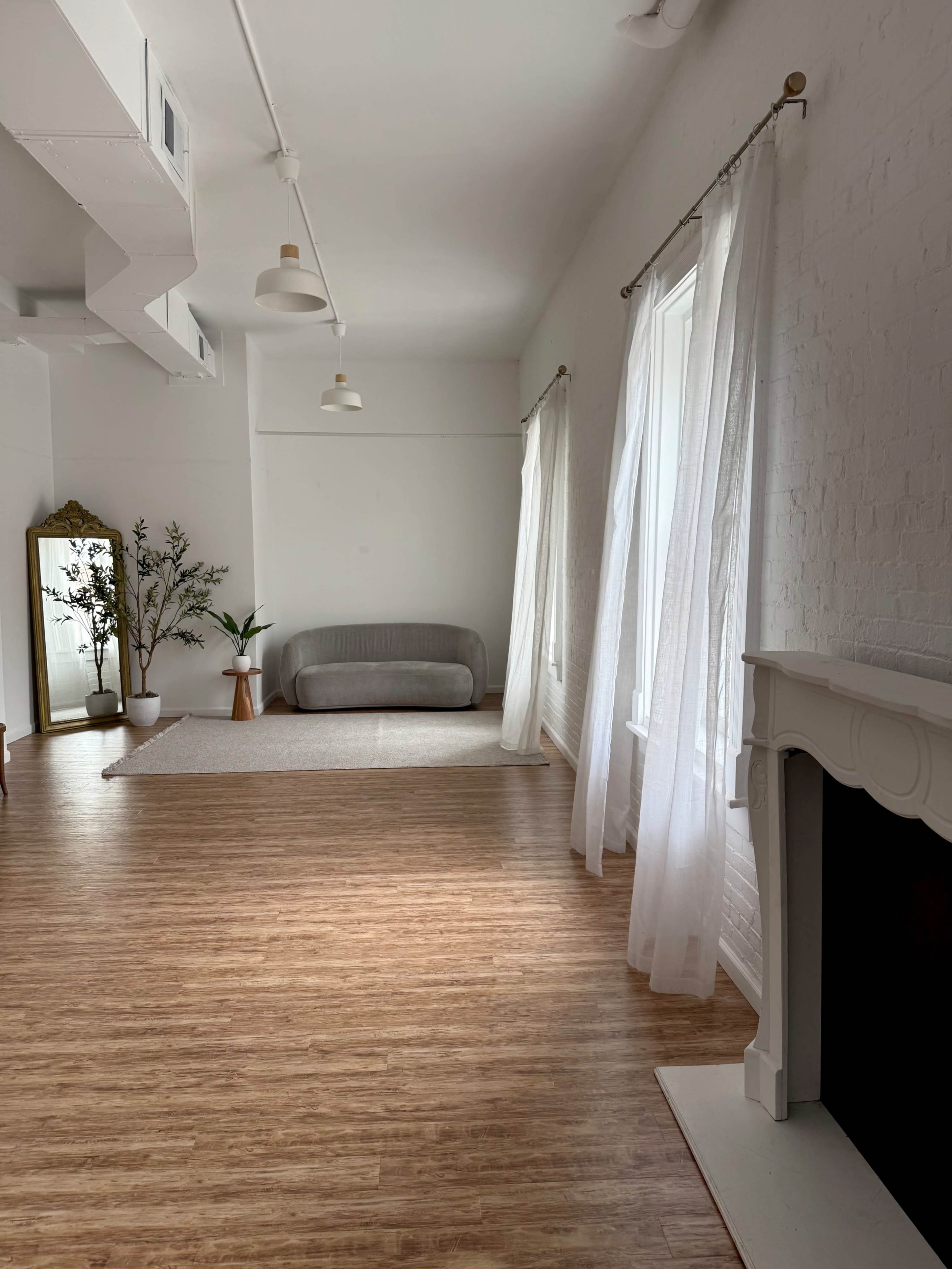 The image shows a bright and airy interior space with a sofa, large windows covered by sheer curtains, a decorative mirror, and minimal furniture.