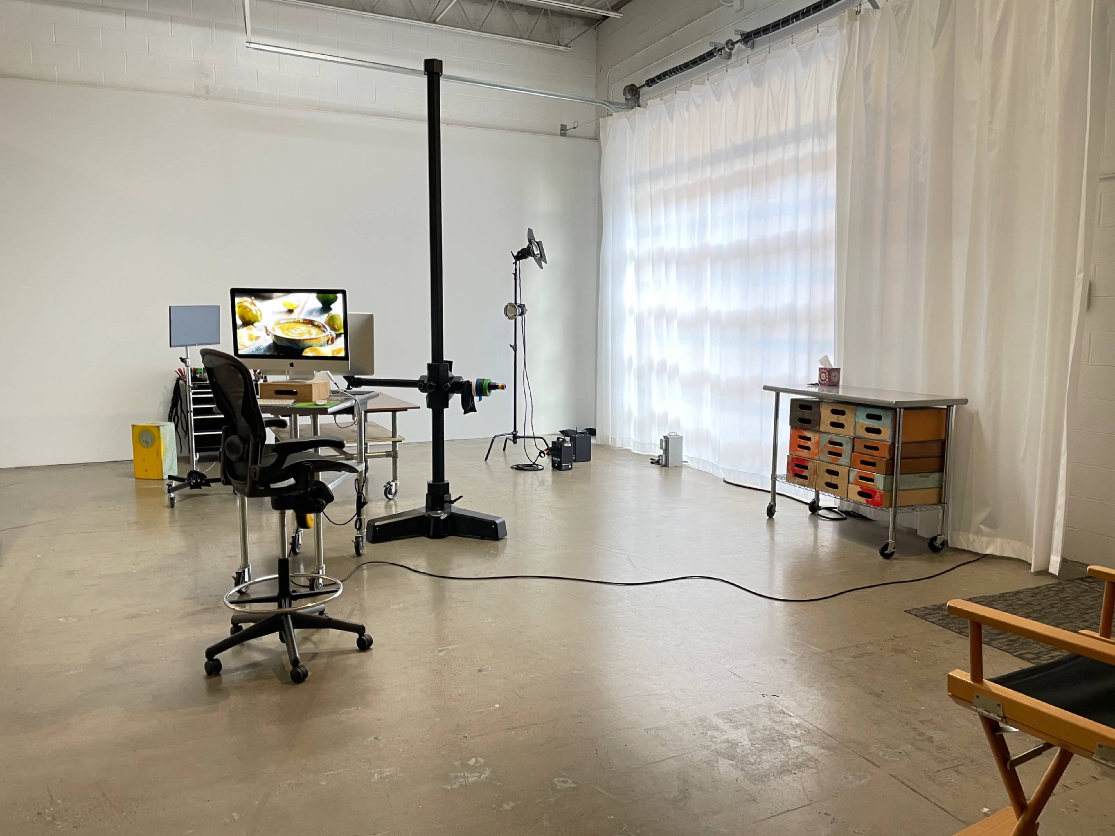 The image shows a spacious studio with a desk, a chair, a monitor displaying food, and a photography light setup, all against a white backdrop.