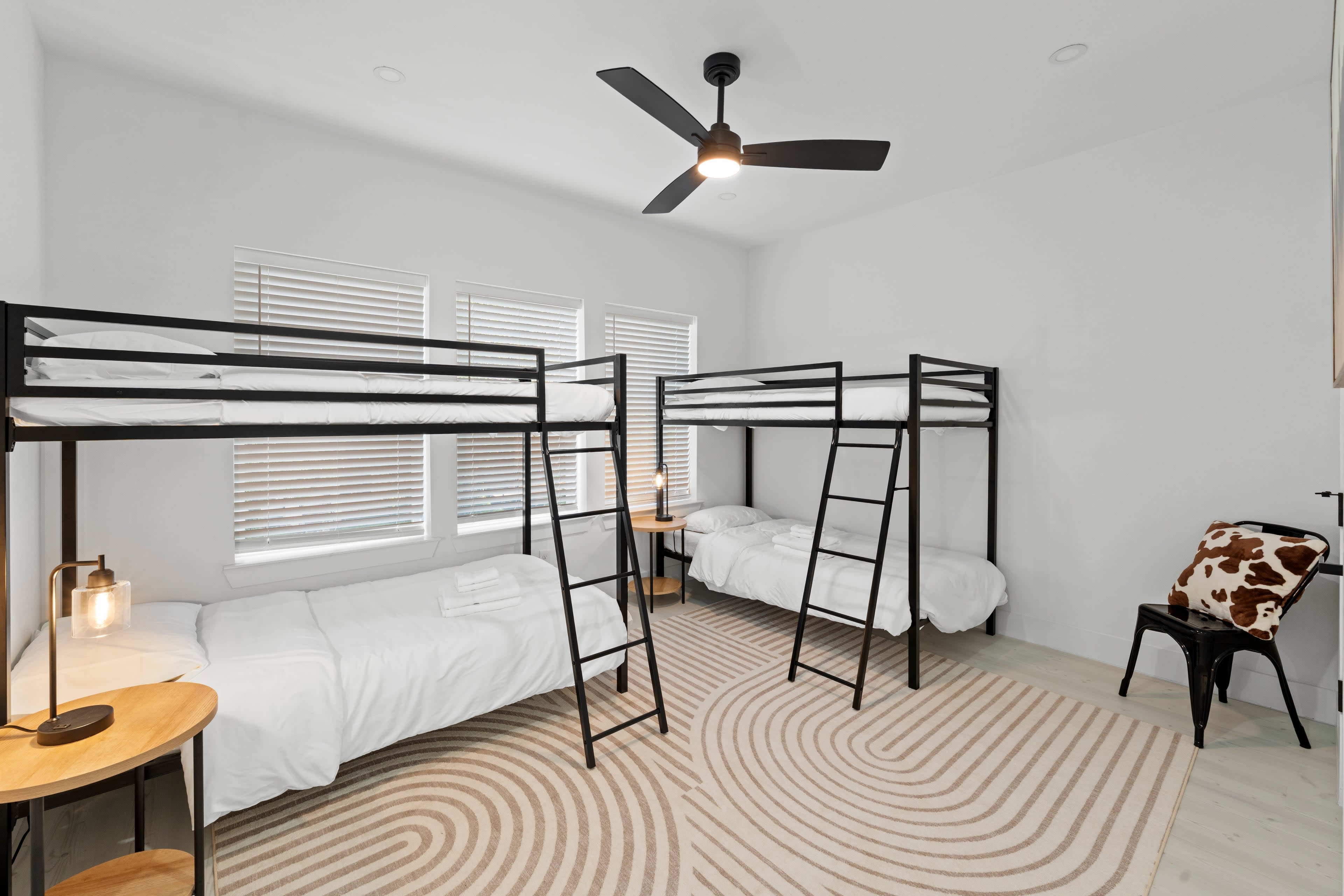 The image shows a modern bedroom with two sets of black metal bunk beds, a single bed, a bedside table with a lamp, and a patterned area rug on the light-colored floor.