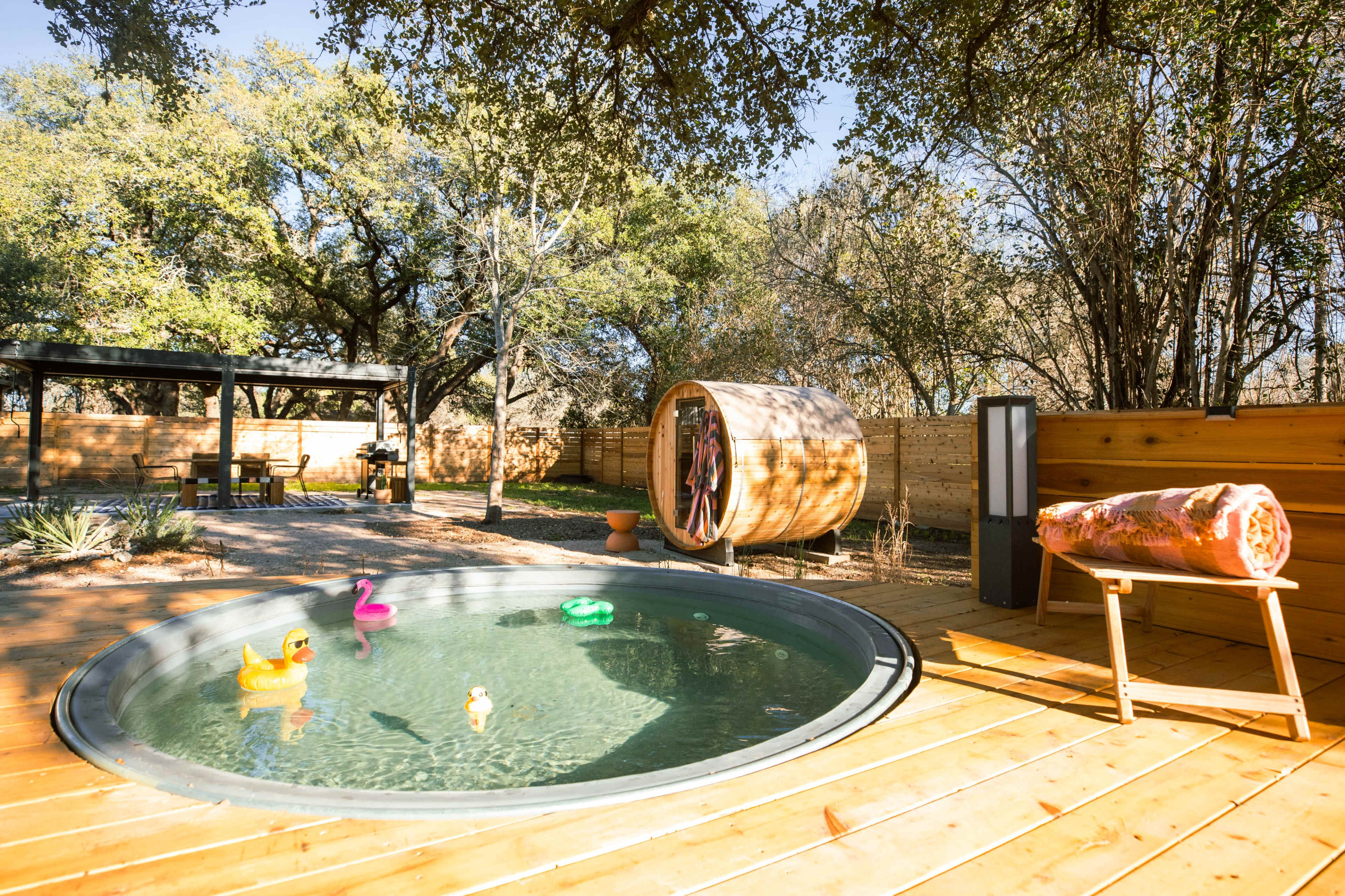 The scene features a circular hot tub surrounded by wooden decking, with colorful rubber ducks floating in the water and a large wooden barrel nearby.
