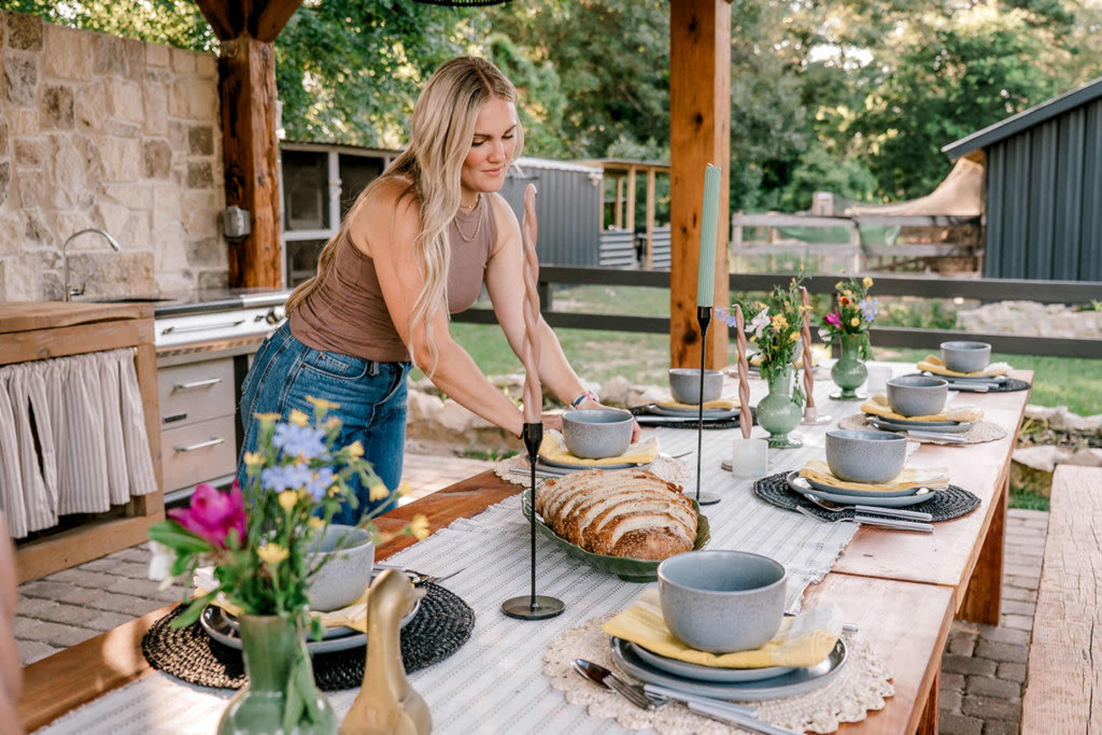 A woman sets a large outdoor dining table with dishes, cutlery, and a loaf of bread in a scenic backyard.