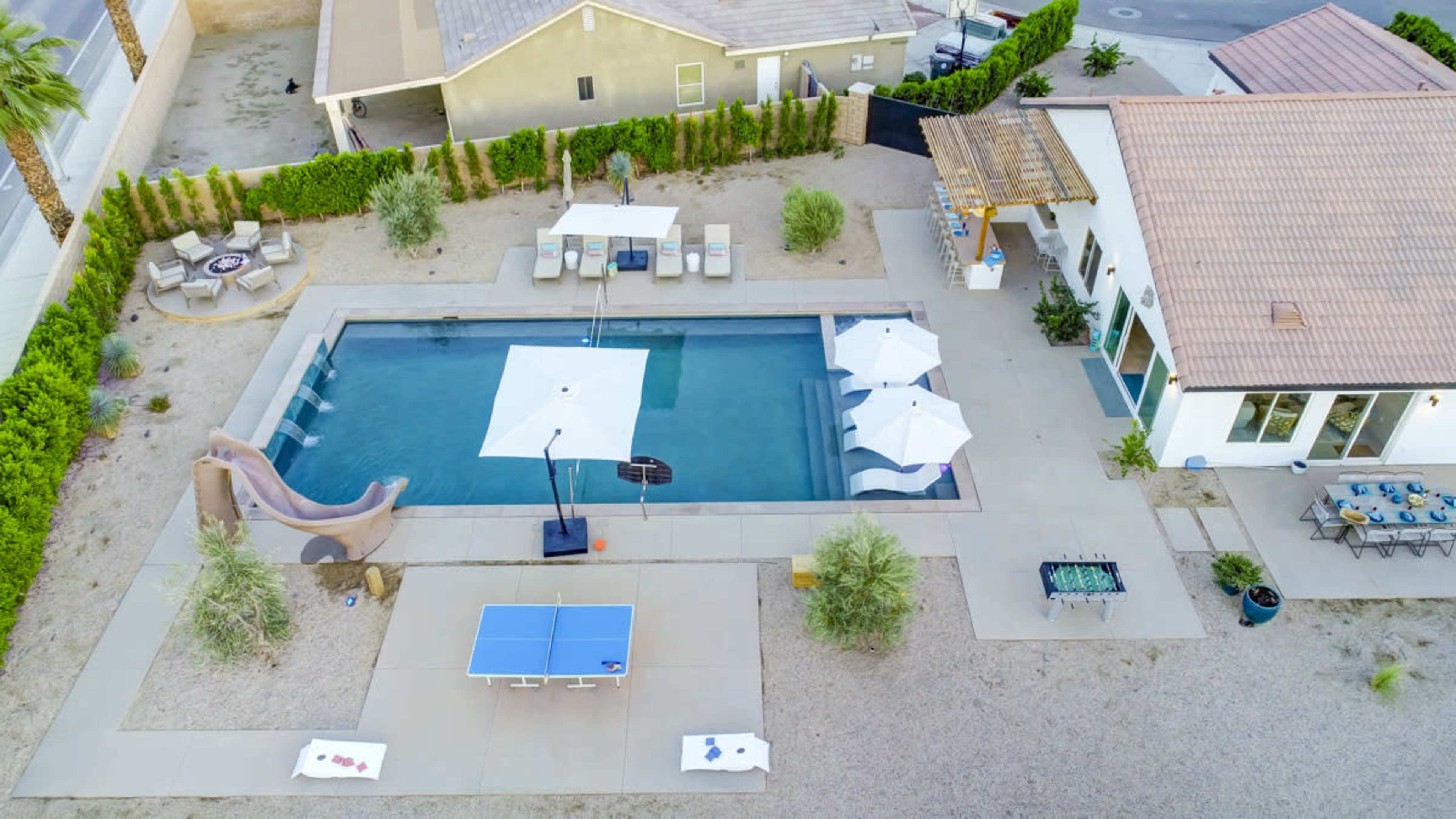 The image shows an aerial view of a landscaped backyard featuring a swimming pool, lounge chairs, a slide, a table tennis setup, and patio umbrellas.