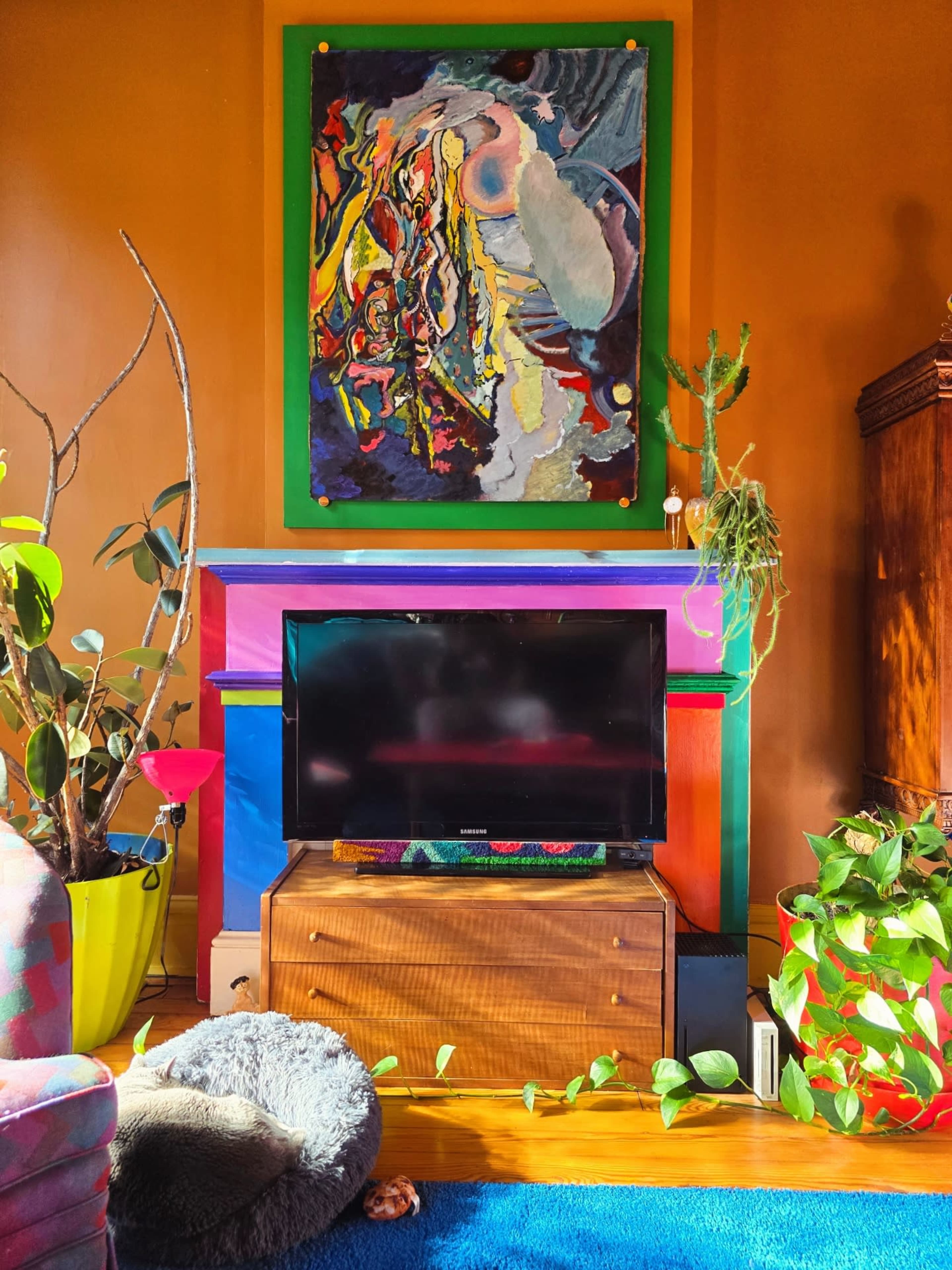 The image features a colorful living room with a television on a wooden cabinet beneath an abstract painting, surrounded by various indoor plants.
